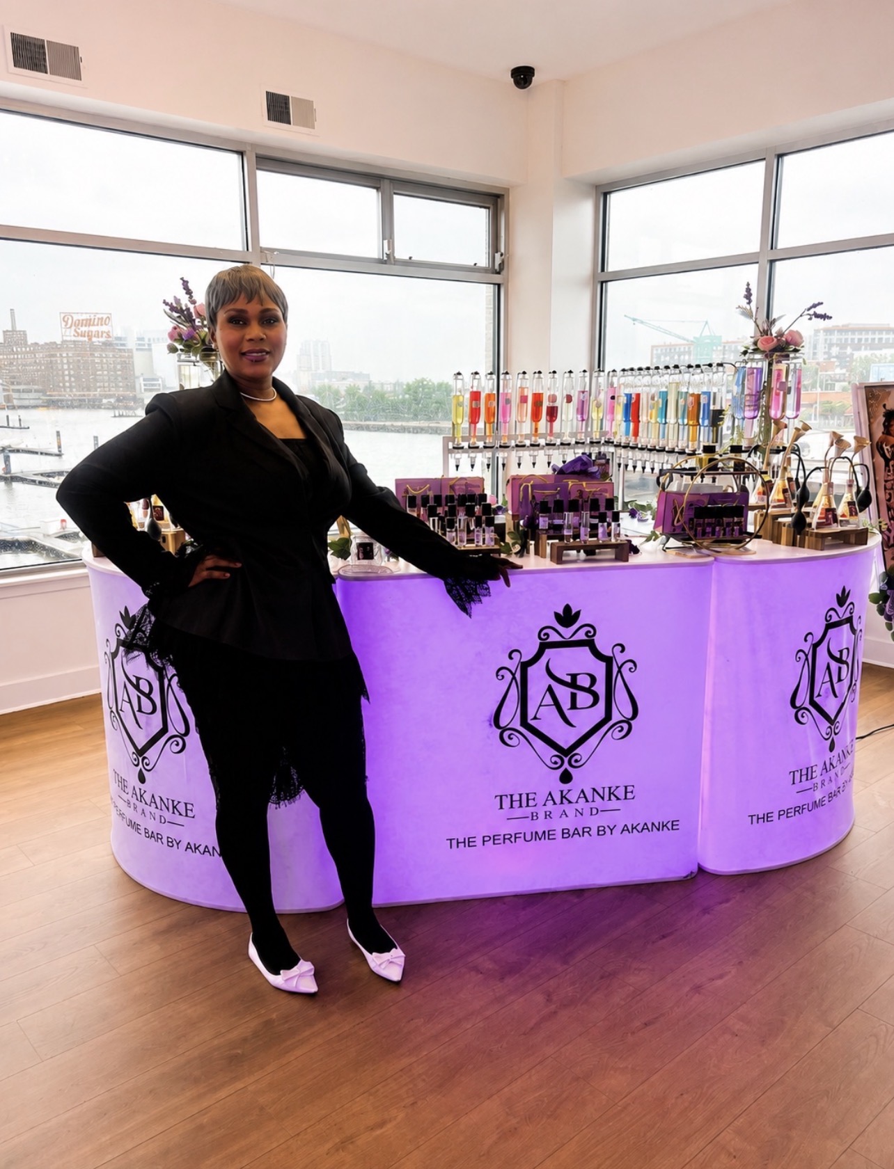 Woman standing next to a jewelry display table with a purple illuminated front, large windows in the background, and cityscape view.