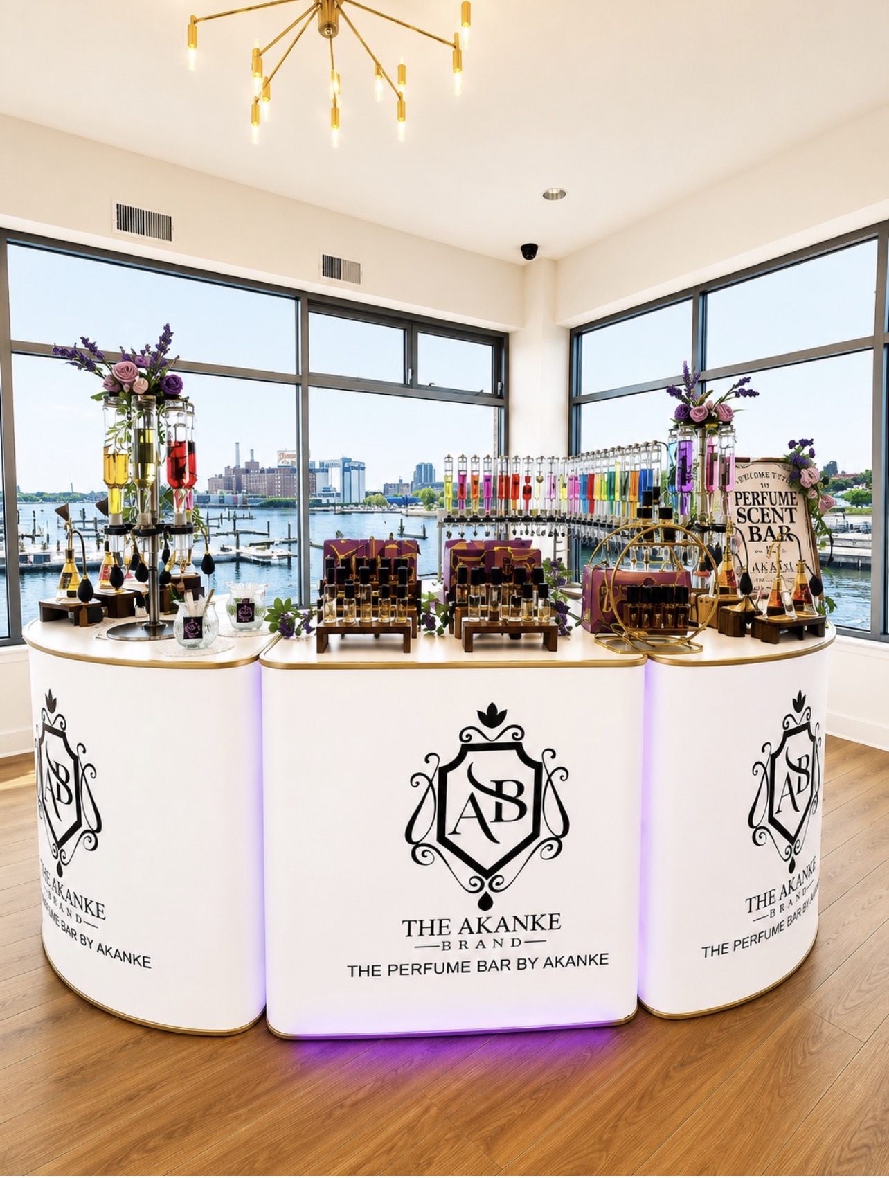 Curved display table with perfume bottles, flowers, and decorative items in front of large windows overlooking a cityscape.