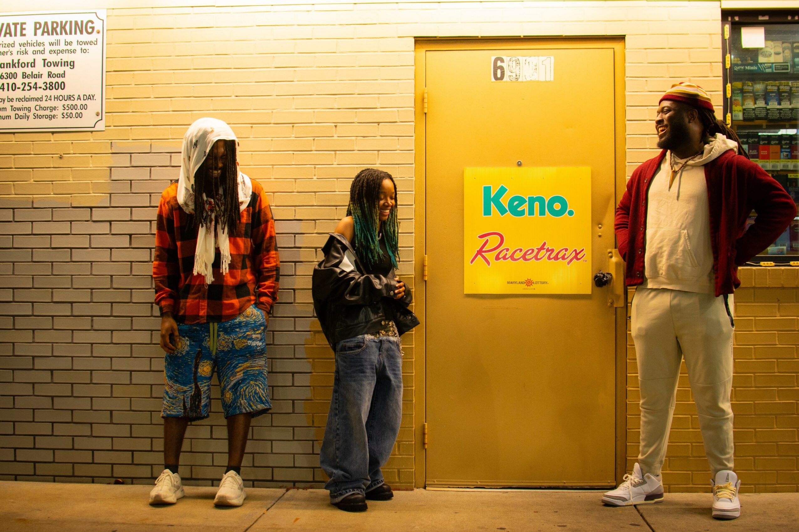 Three people stand outside a yellow door with a Keno sign, against a brick wall, in daytime. Word count: 24.