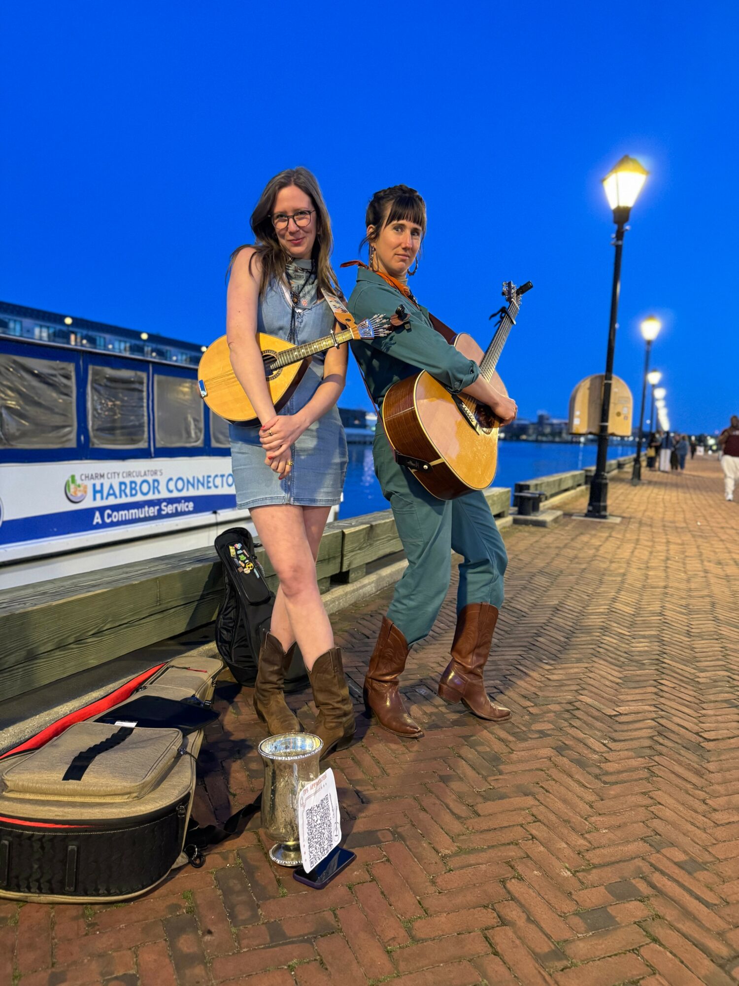 Two women with guitars perform on a brick walkway near a harbor, with a boat and streetlights in the background.