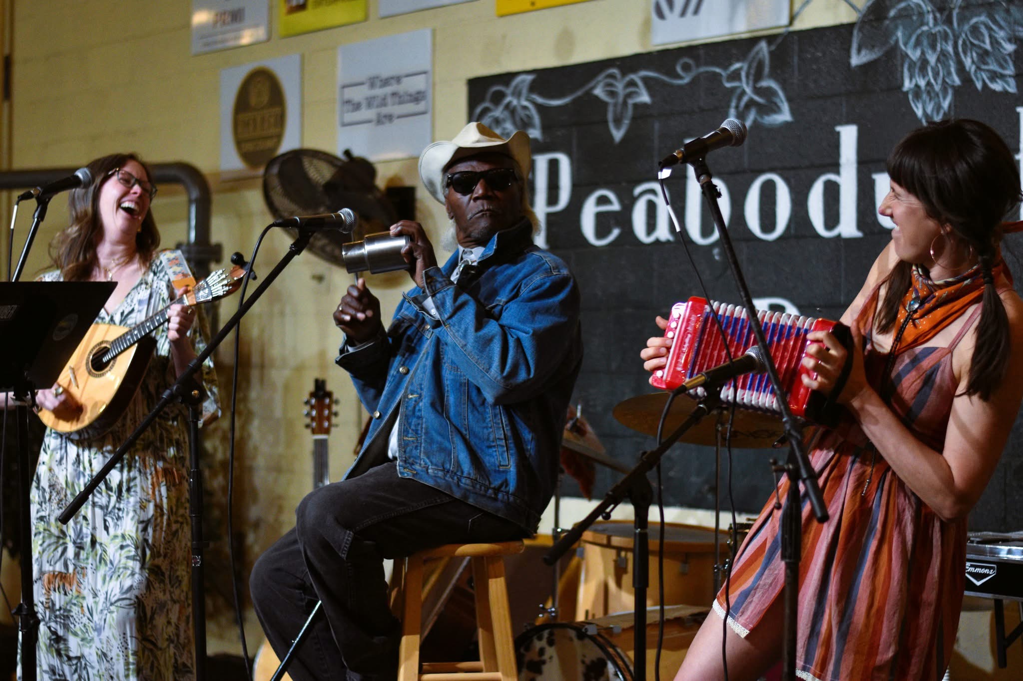 Three musicians perform on stage, one playing guitar, one singing, and one playing a percussion instrument, with a blackboard and posters behind.