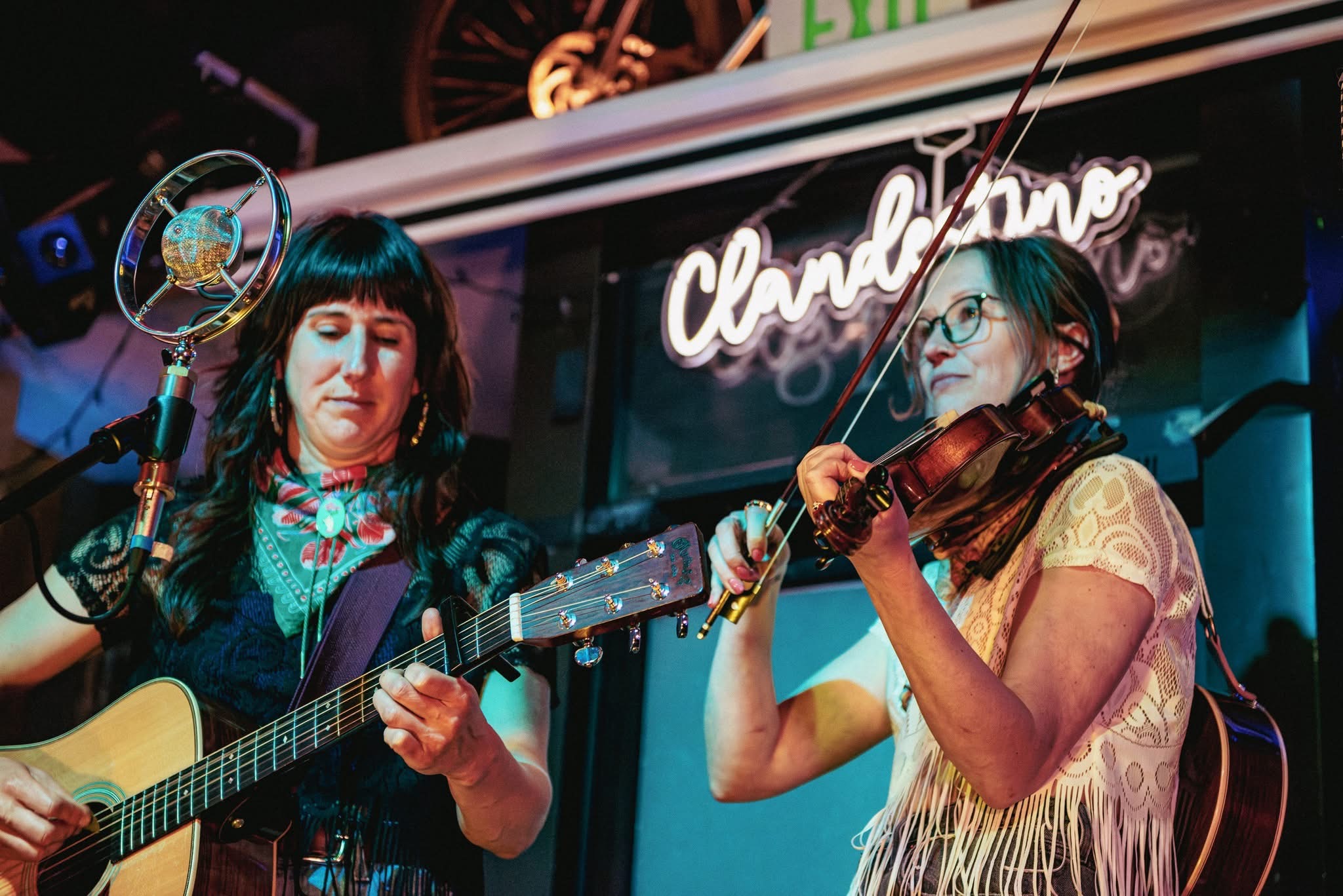 Two women perform music on stage, one playing guitar and the other playing violin, with a neon sign in the background.