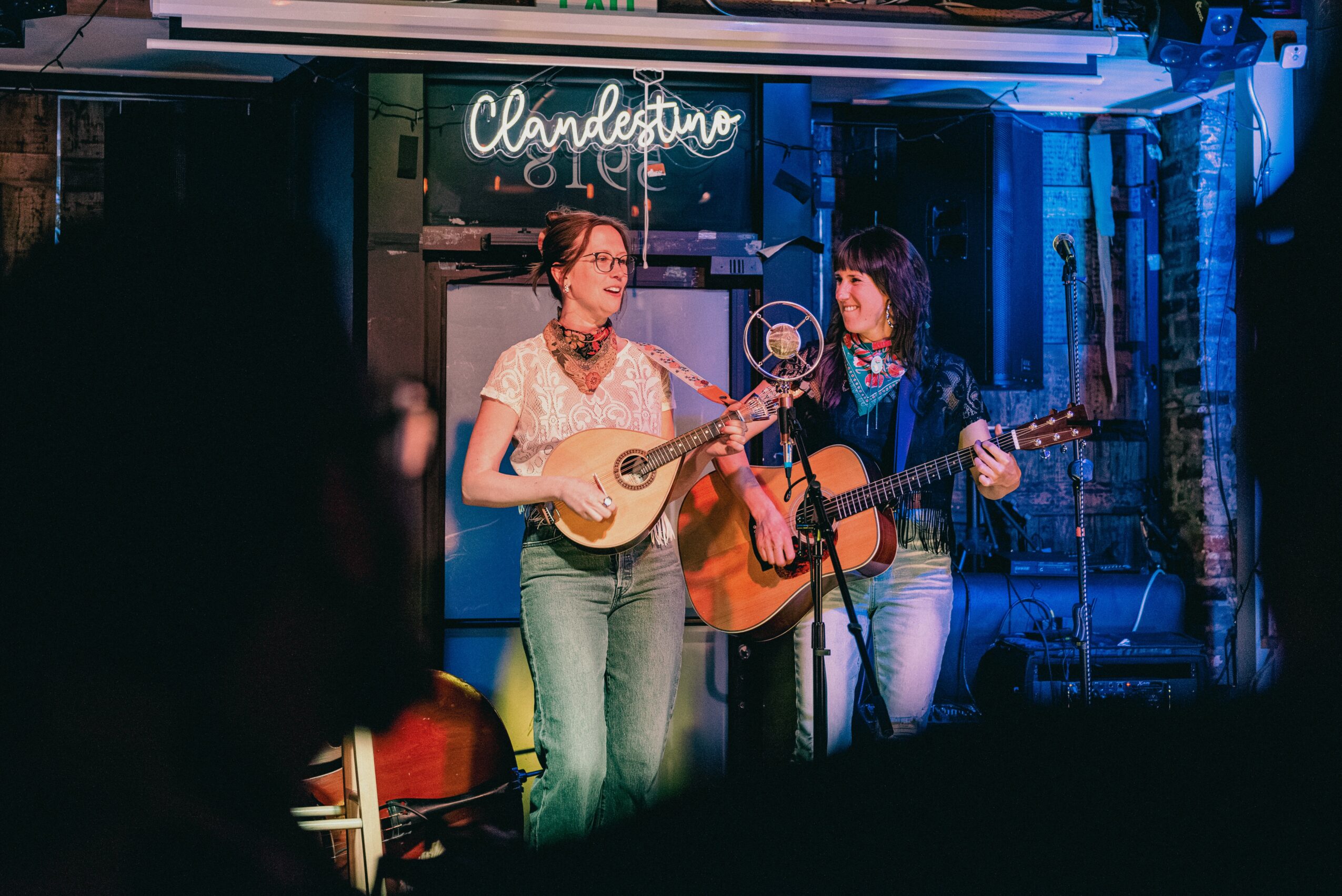 Two women perform with guitars on stage in a dimly lit venue, neon sign reads 'Clandestino Lounge'.