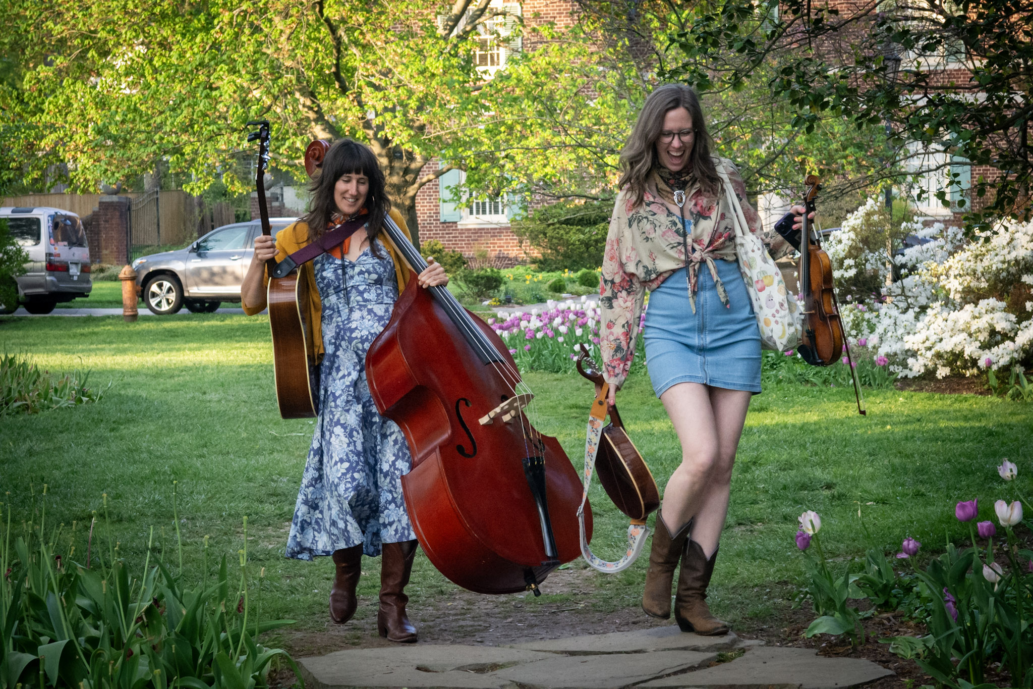 Two women walking in a park with musical instruments, one with a guitar and a large red double bass, surrounded by greenery and flowers.