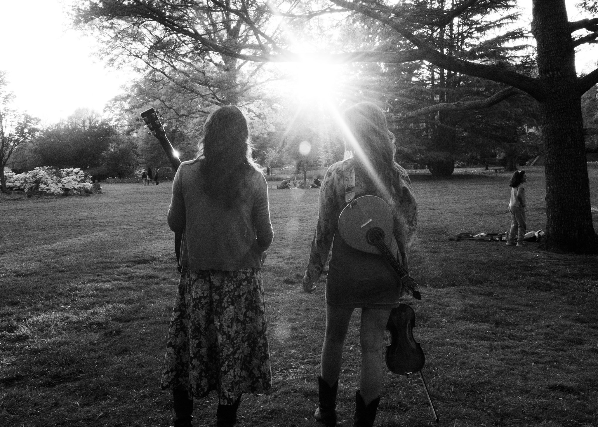 Two people with guitars walking in a park with trees and sunlight shining through, other people in the background.