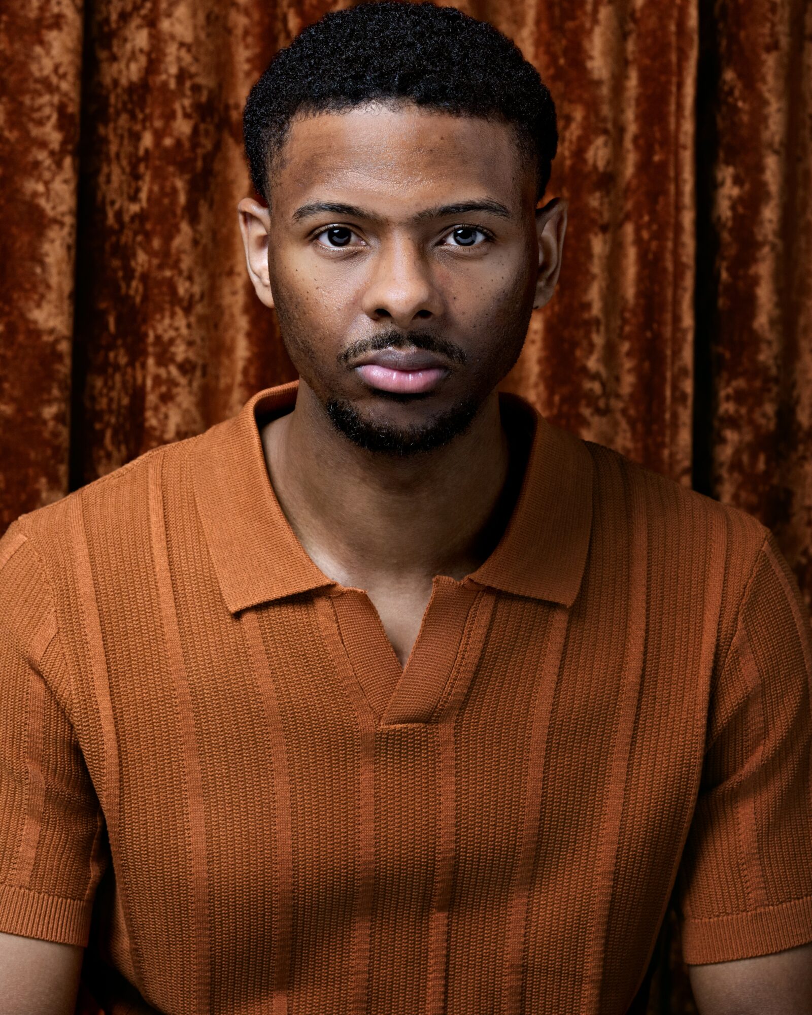 Young man with short curly hair and a serious expression, wearing a brown polo shirt, standing in front of a brown curtain.