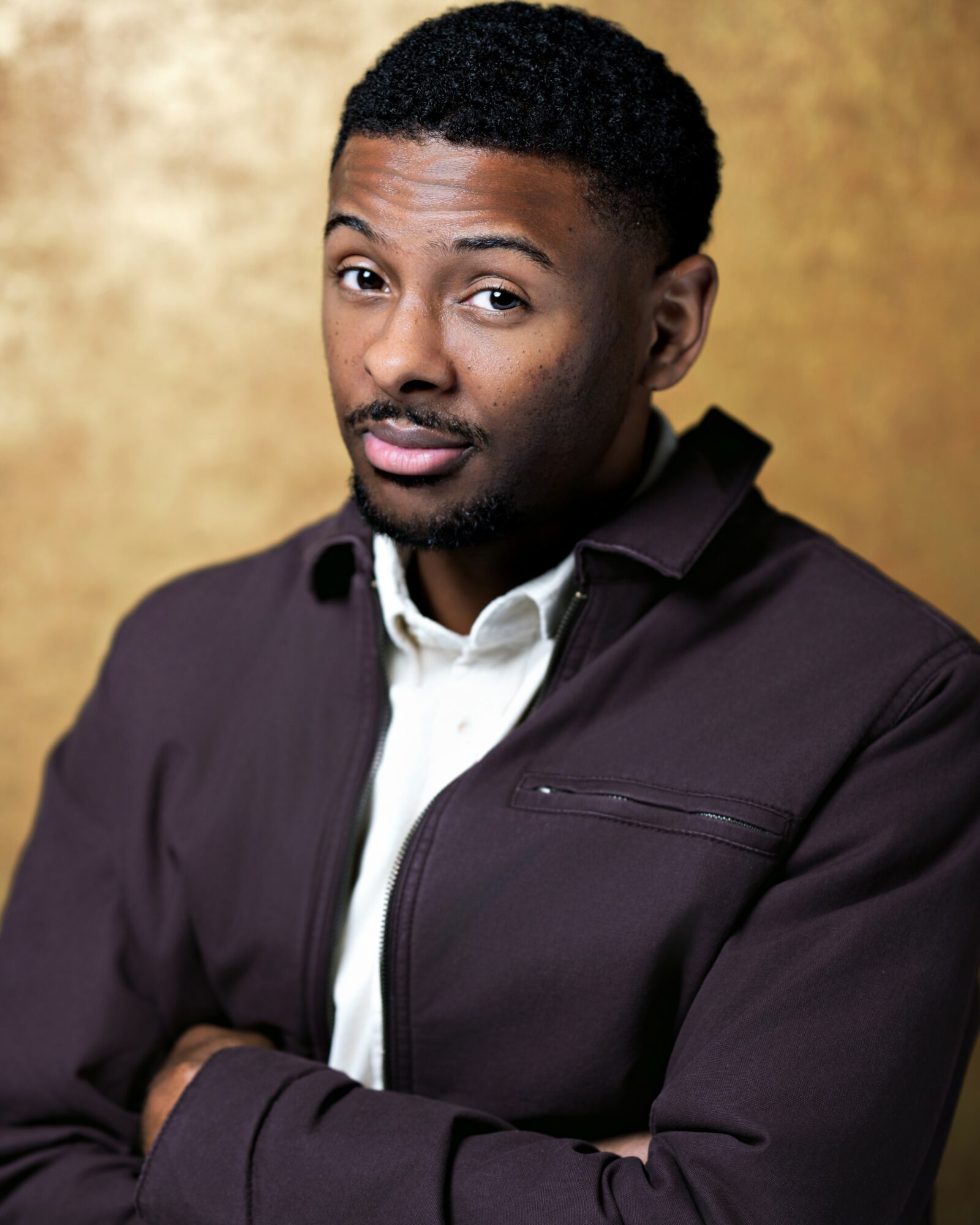 Man with short curly hair and a beard, wearing a dark jacket over a white shirt, standing against a gold background.