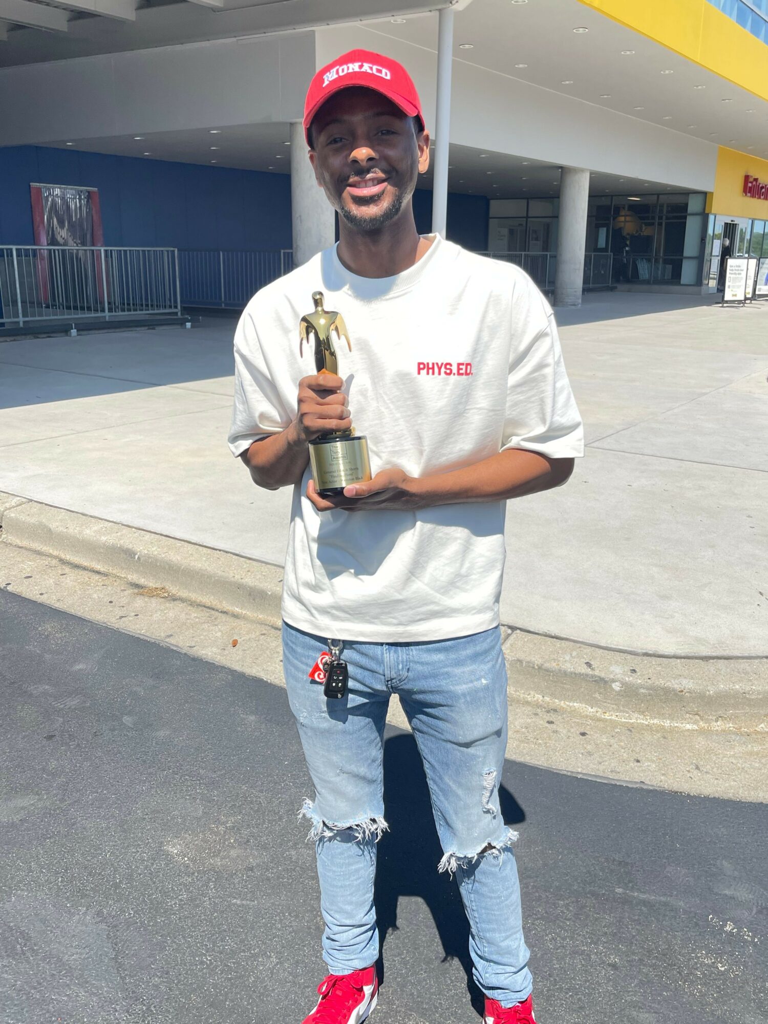 Young man with a beard wearing a red cap, white t-shirt, ripped jeans, and red shoes, holding a trophy.