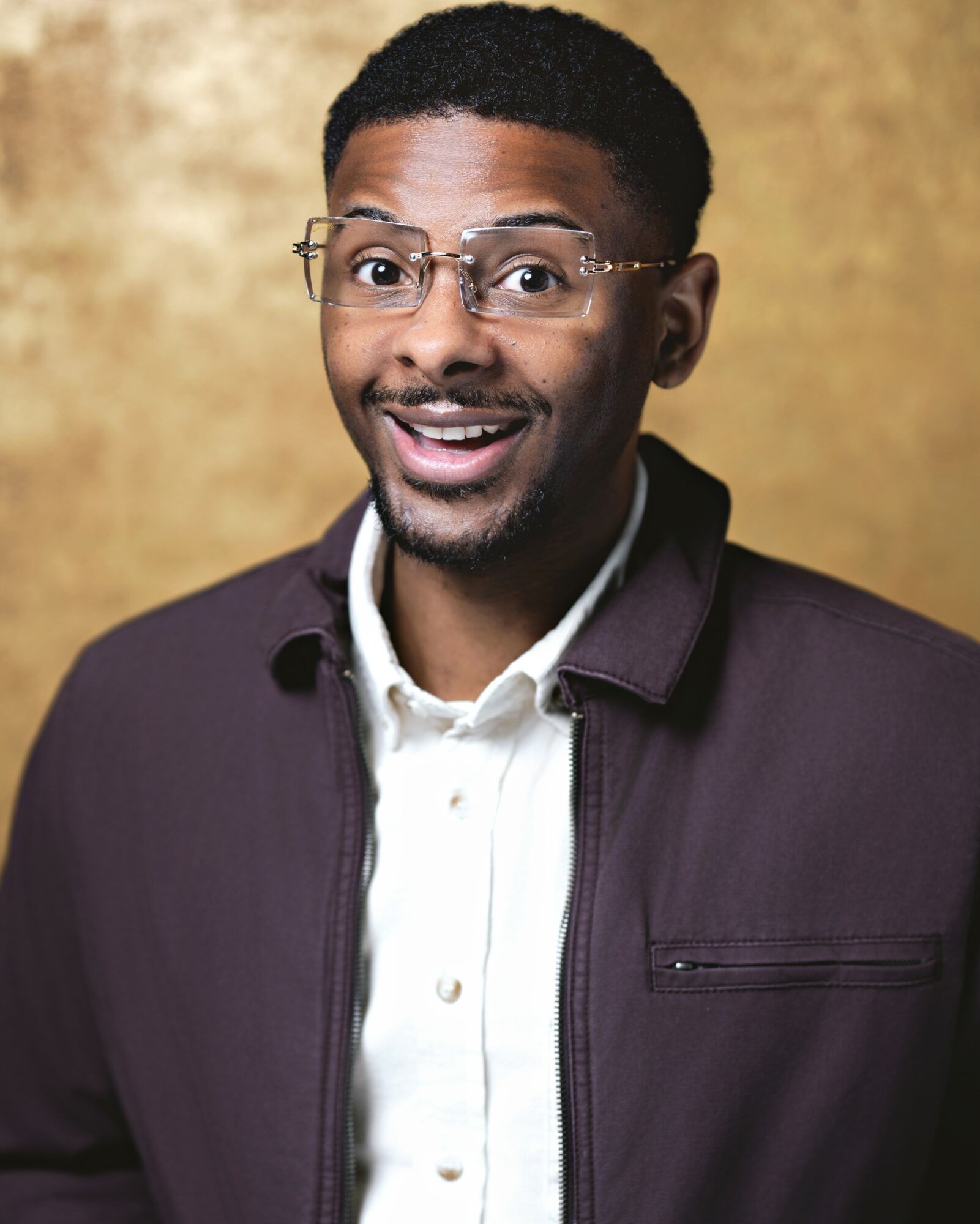 Man with glasses smiling, wearing a dark jacket and white shirt, against a gold background.