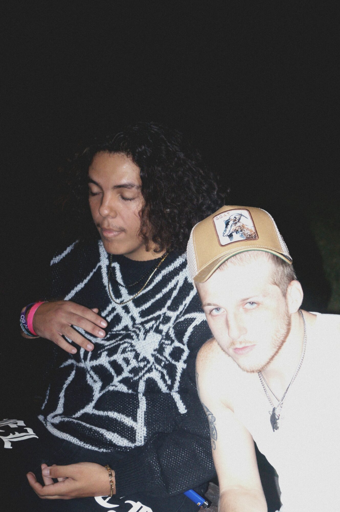 Two people, one with curly hair and a patterned sweater, and another with a cap, are close together in a dark setting.
