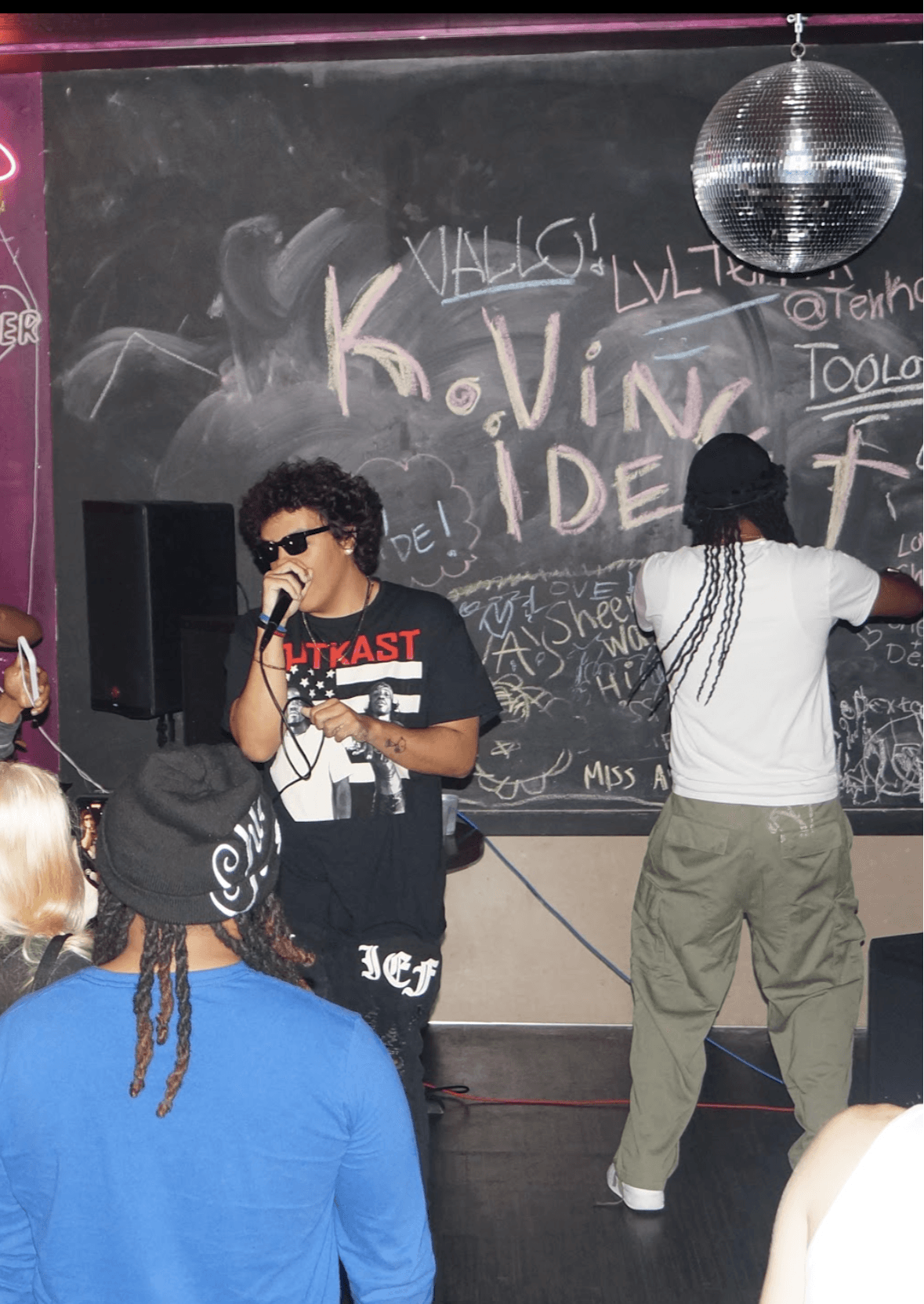 Two people on stage, one with sunglasses and a microphone, the other with long braids, in front of a chalkboard with writing and a disco ball.