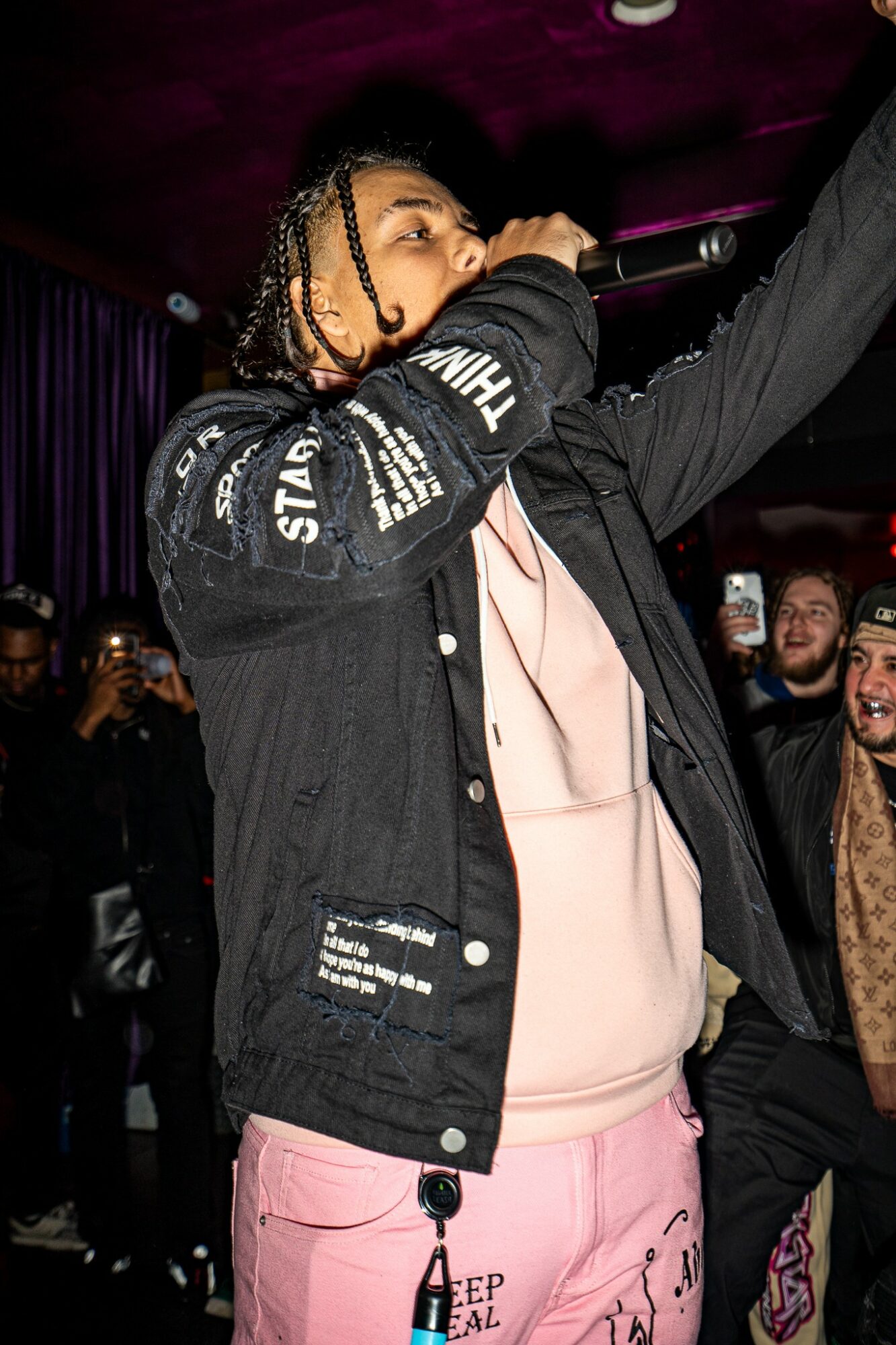 Performer with braided hair singing into a microphone on stage, wearing a black jacket and pink pants, audience in background.