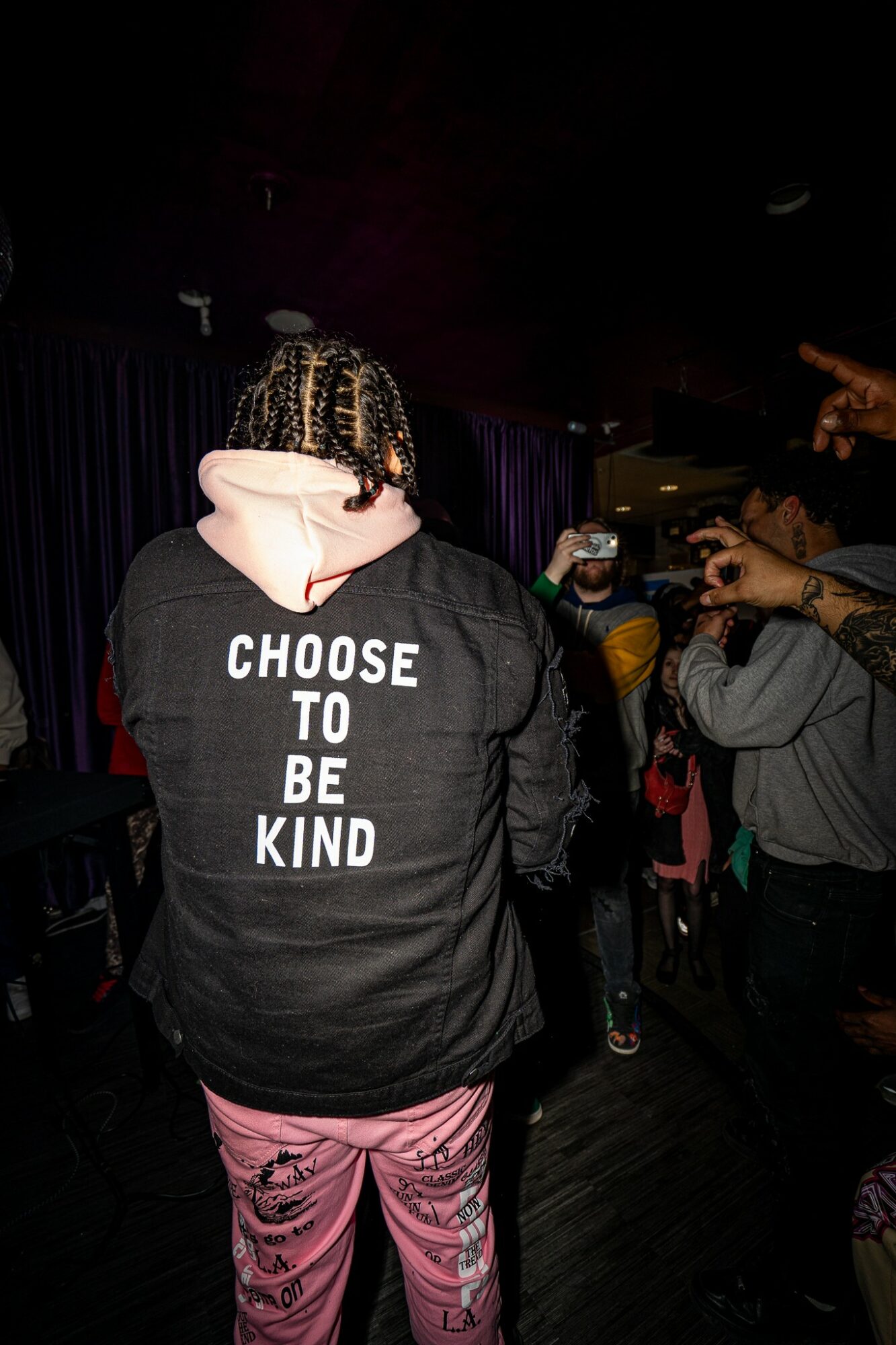Person with braided hair wearing a black hoodie with white text, pink pants, in a dark room with other people.