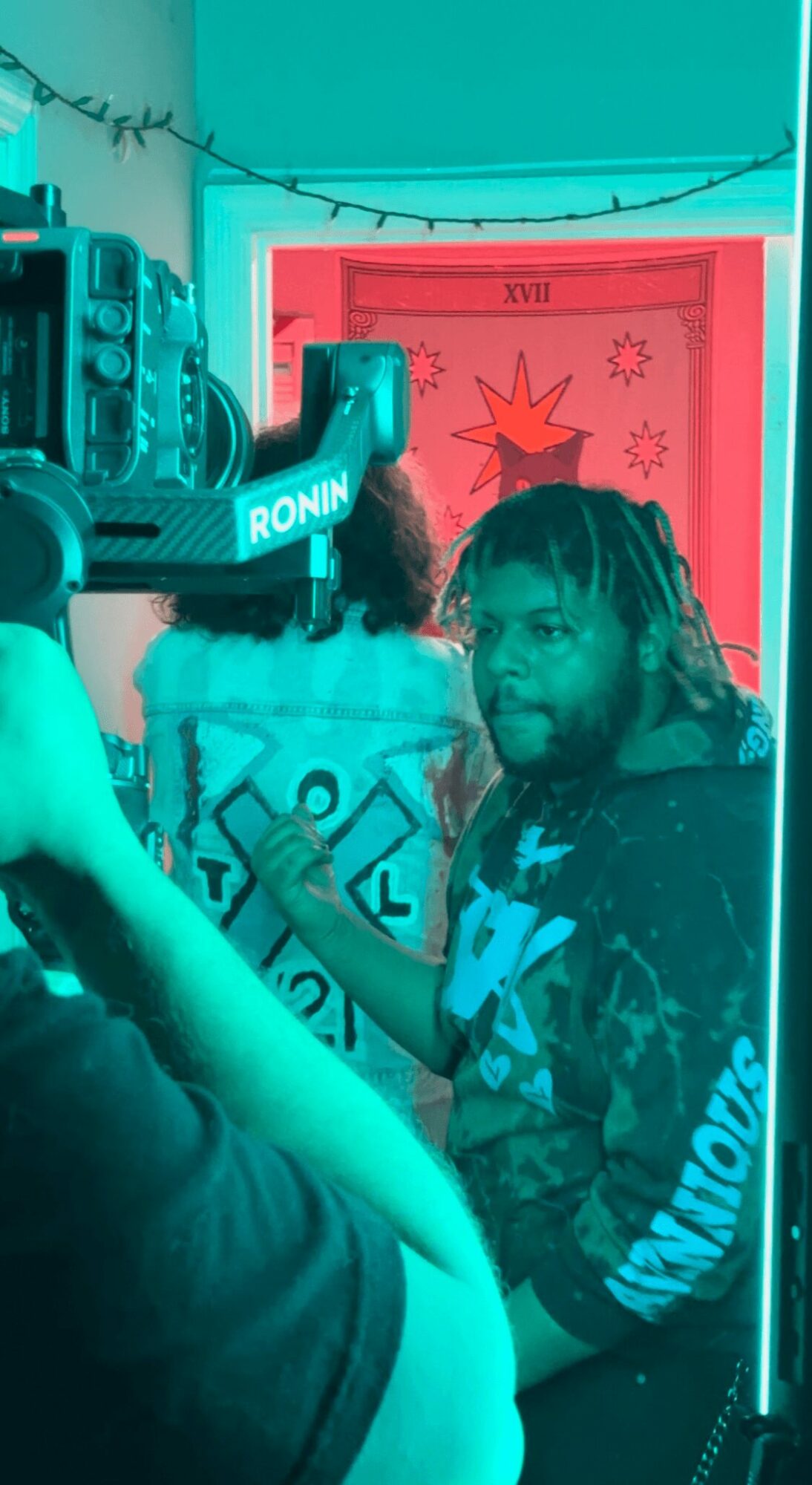 Two people in a room with a red framed picture, one with dreadlocks, the other with curly hair, and a camera in the foreground.
