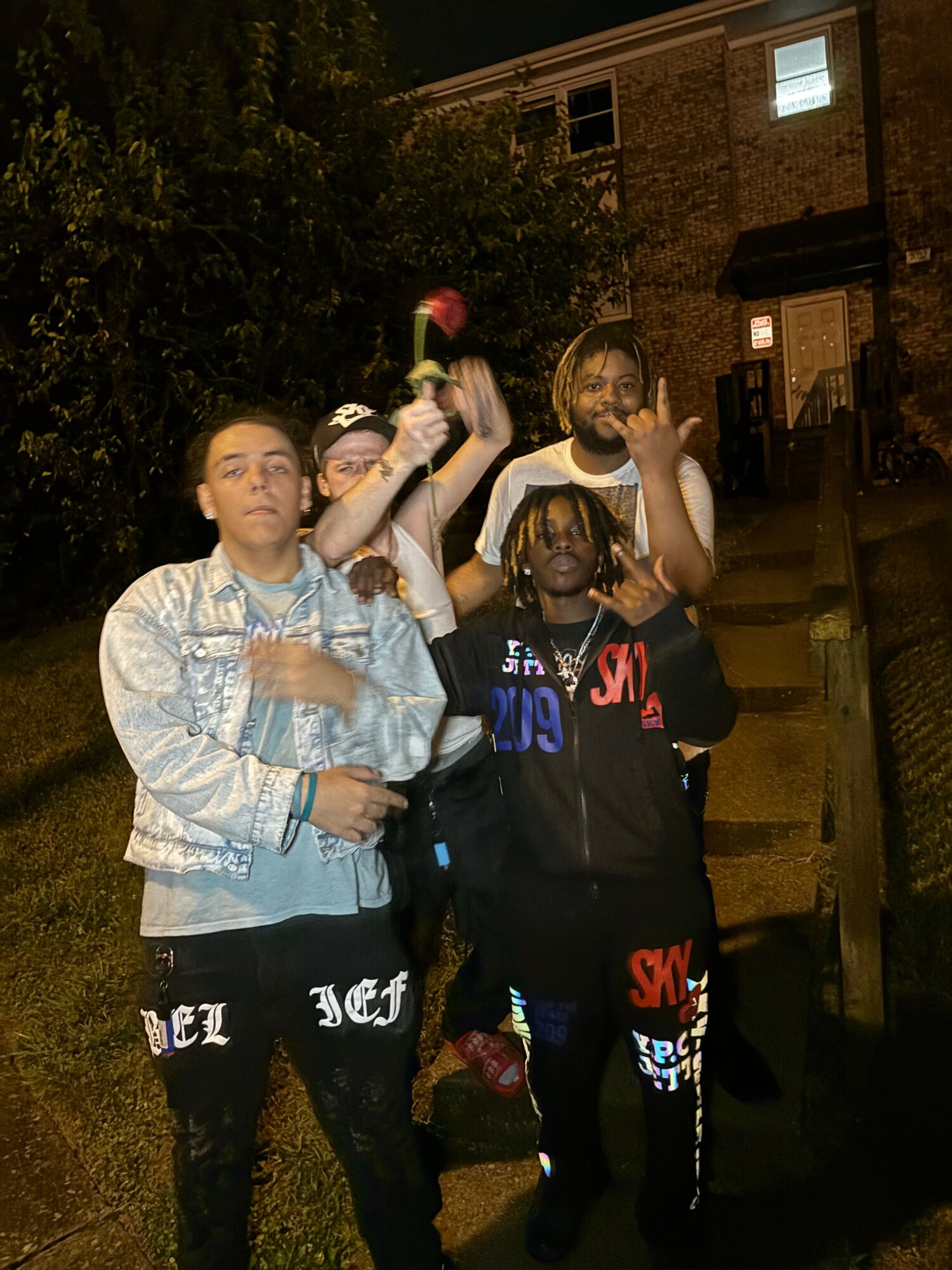 Four people standing outdoors at night, some holding drinks, with trees and buildings in the background.