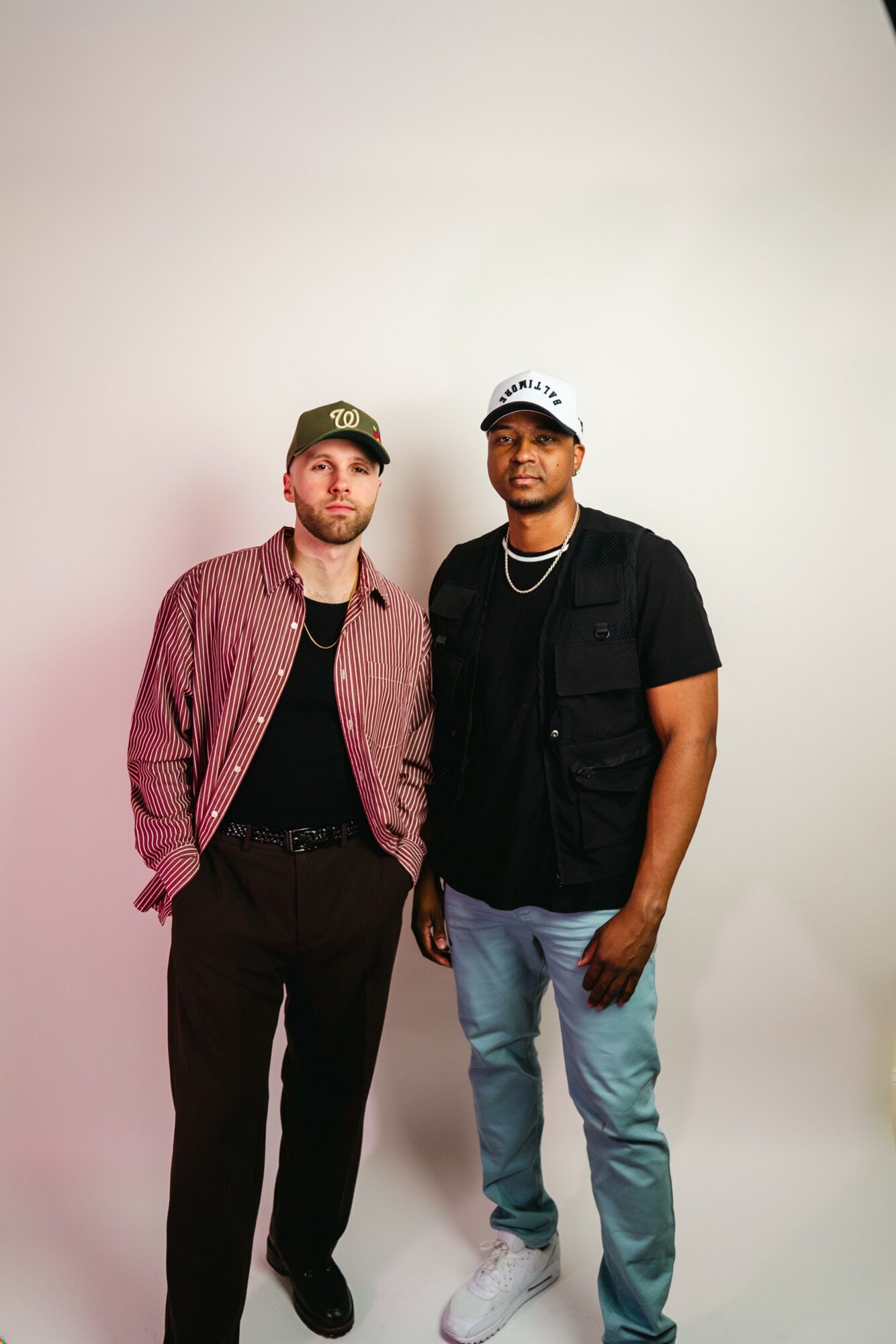 Two men stand side by side against a plain background, one wearing a red jacket and black pants, the other in a black shirt and light blue jeans.