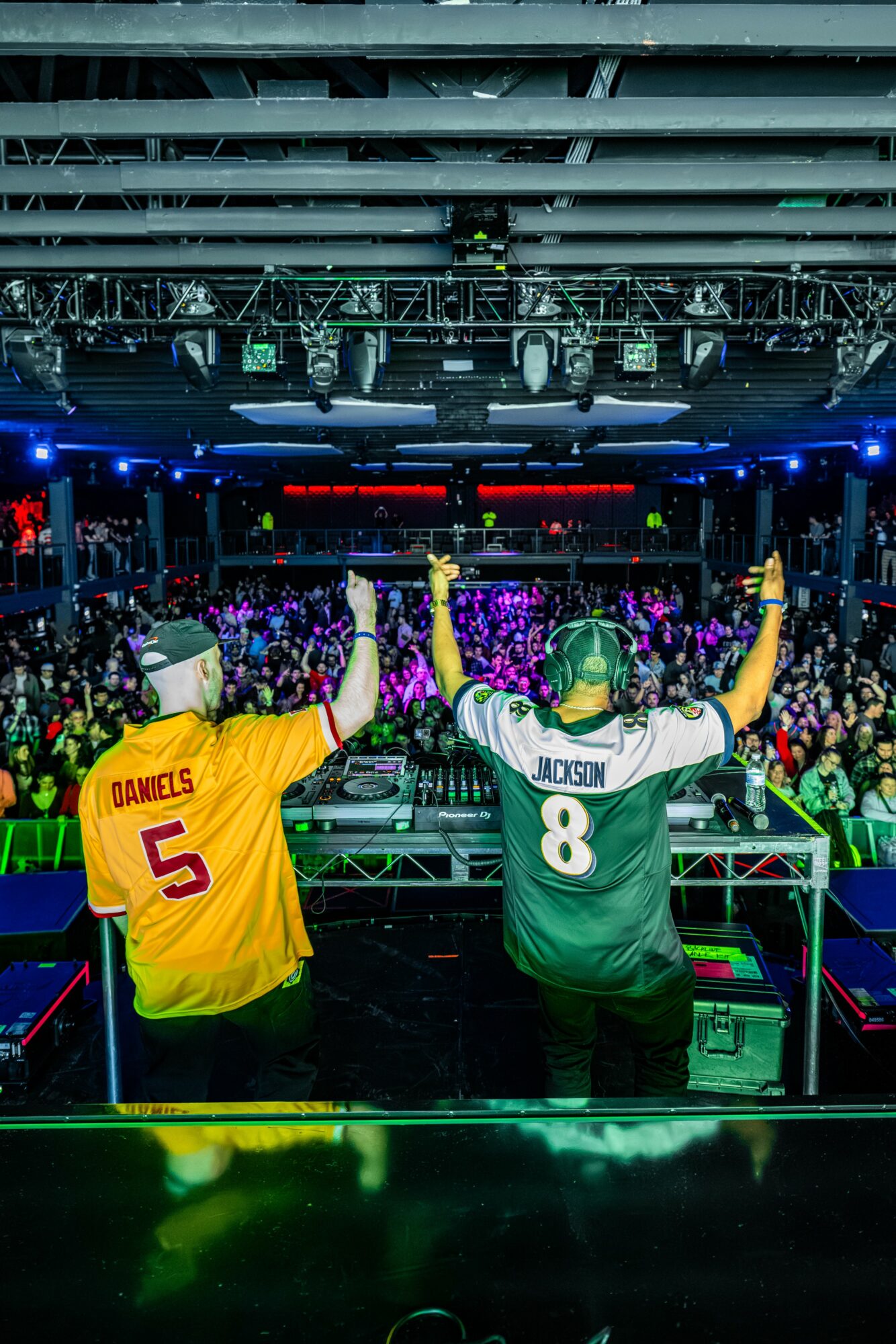 Two DJs perform on stage with a crowd in a large indoor venue, colorful lights, and a high ceiling.