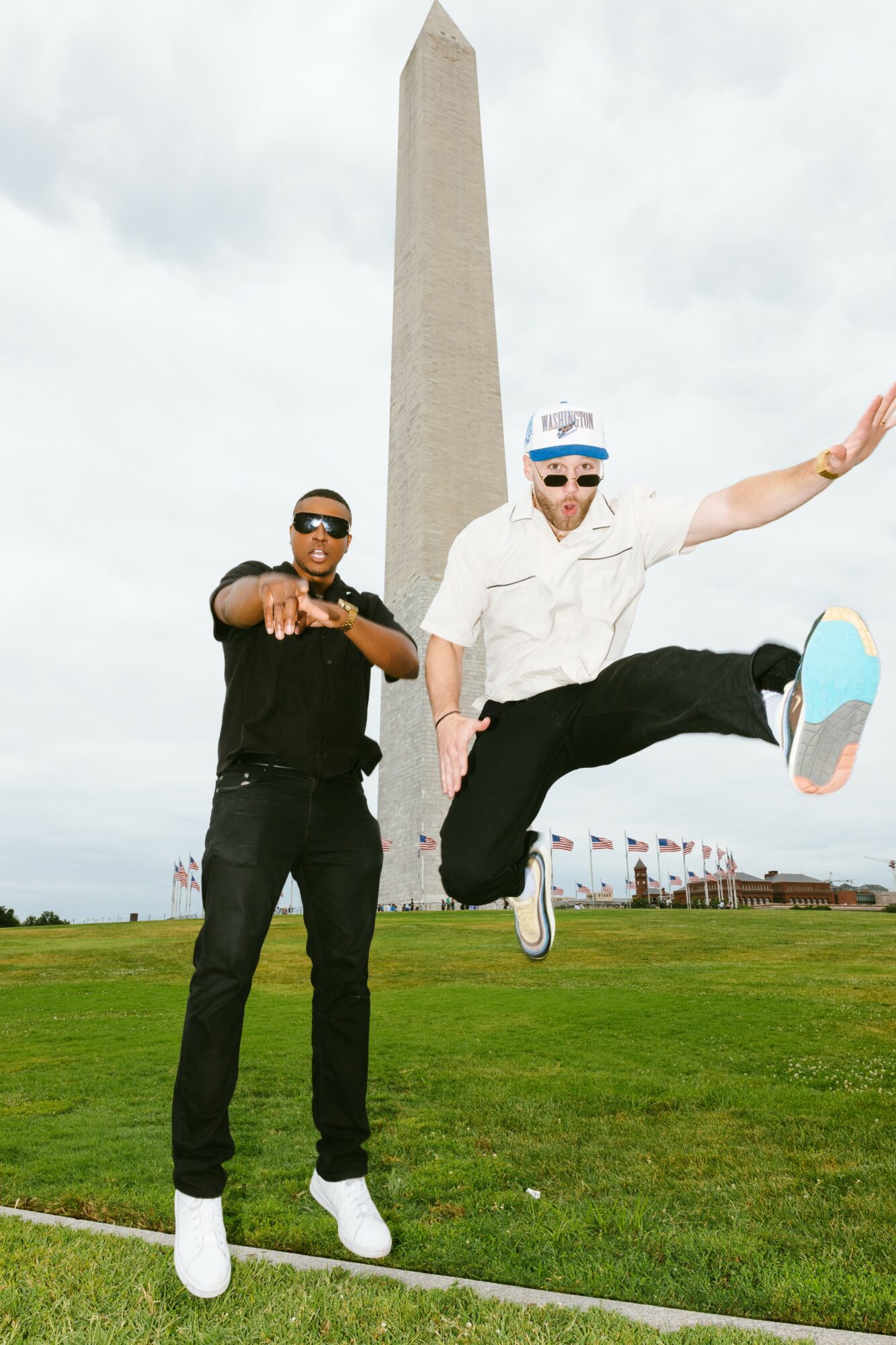 Two men standing on grass near the Washington Monument, one jumping with arms outstretched, both wearing sunglasses and casual clothing.