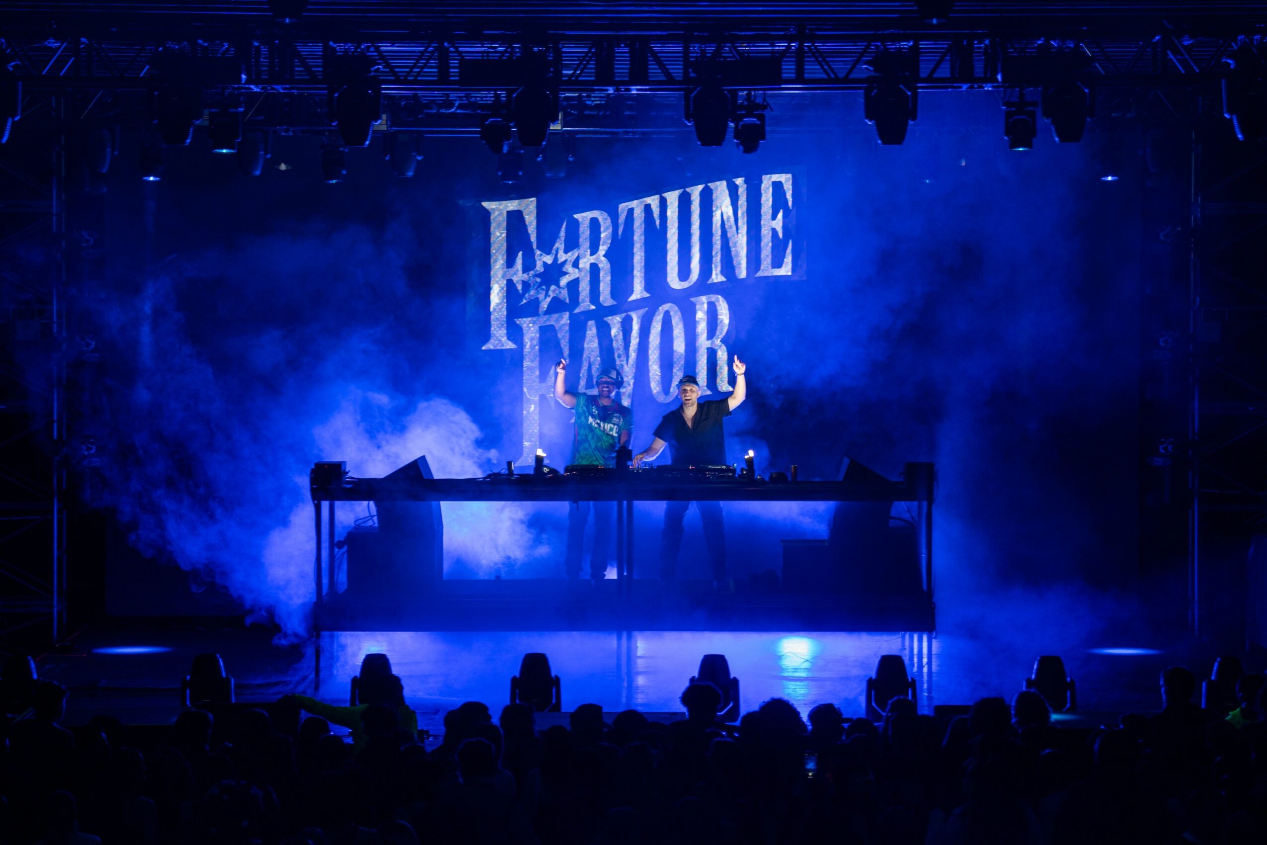 Stage with blue lighting, a DJ booth, and a large screen displaying 'Fortune Favor' in the background, audience silhouettes visible.