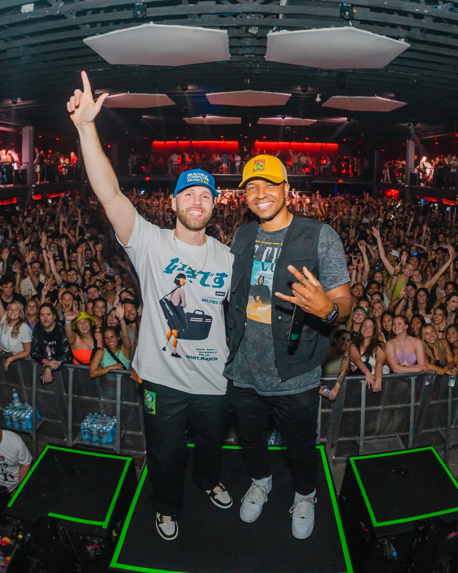 Two men stand on stage with a large crowd behind them, smiling and making hand gestures, in a concert venue.