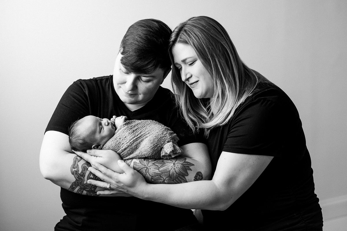 Two adults hold a newborn baby wrapped in a blanket, looking at the baby lovingly, in black and white.