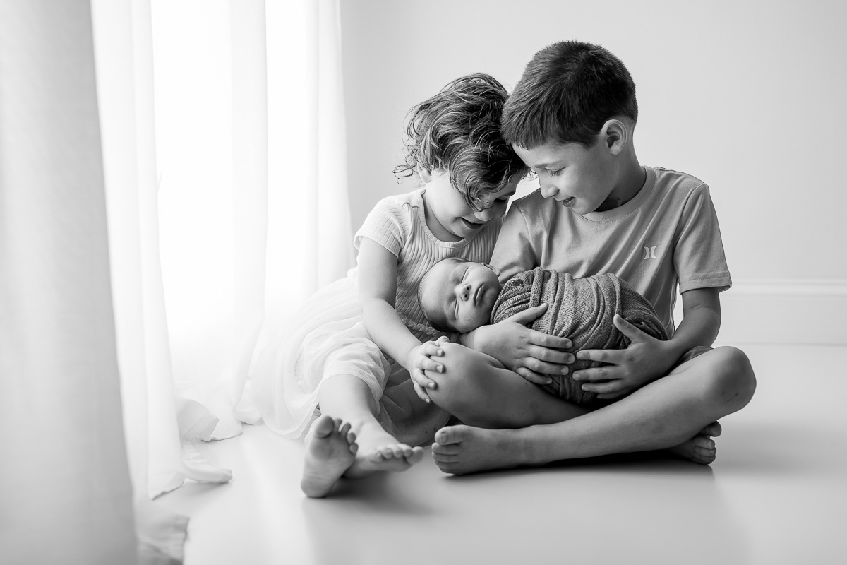 Two children gently hold a newborn baby, sitting on the floor near a window with curtains, in black and white.