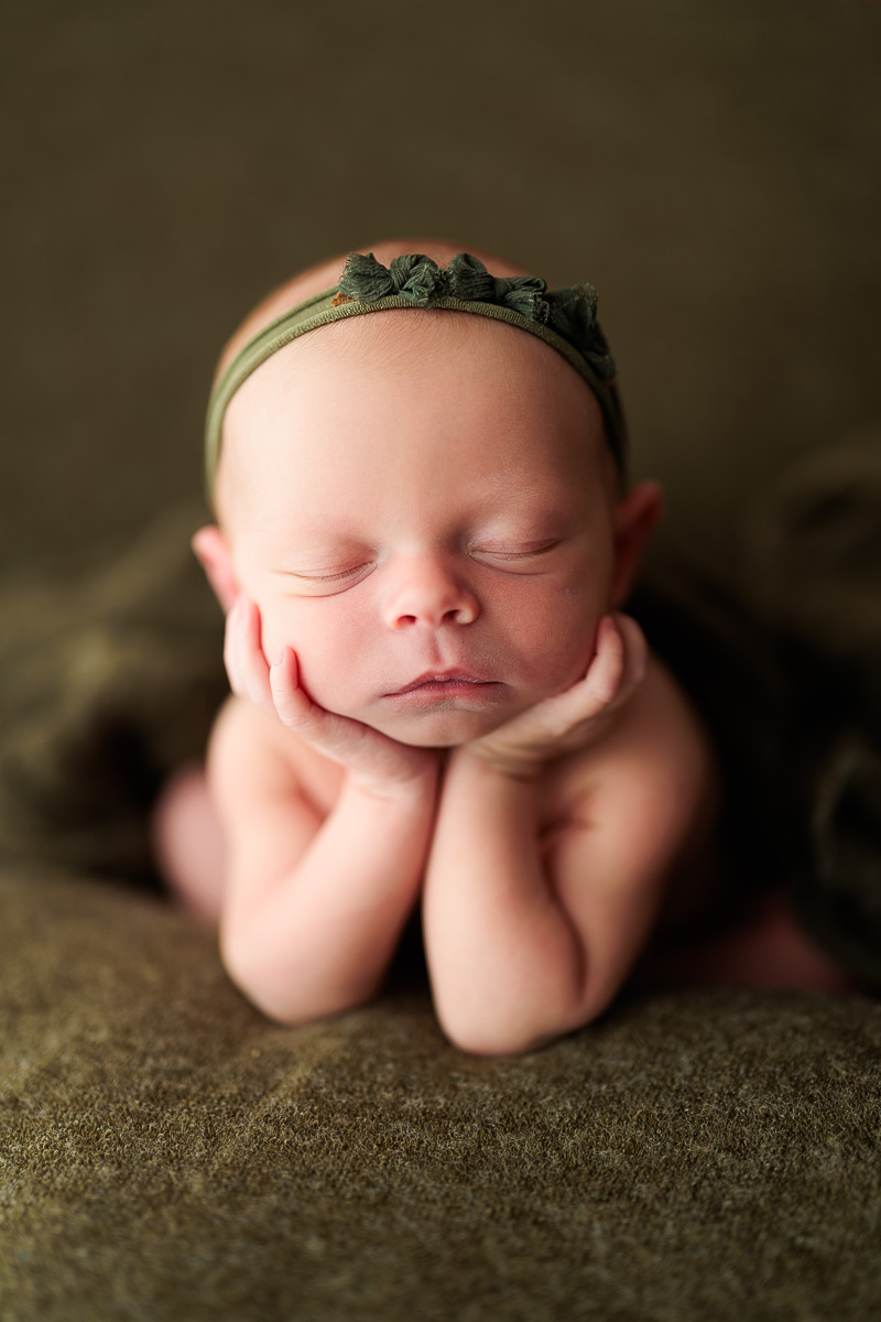 Baby with closed eyes, resting face on hands, wearing a headband with a bow, lying on a soft surface.