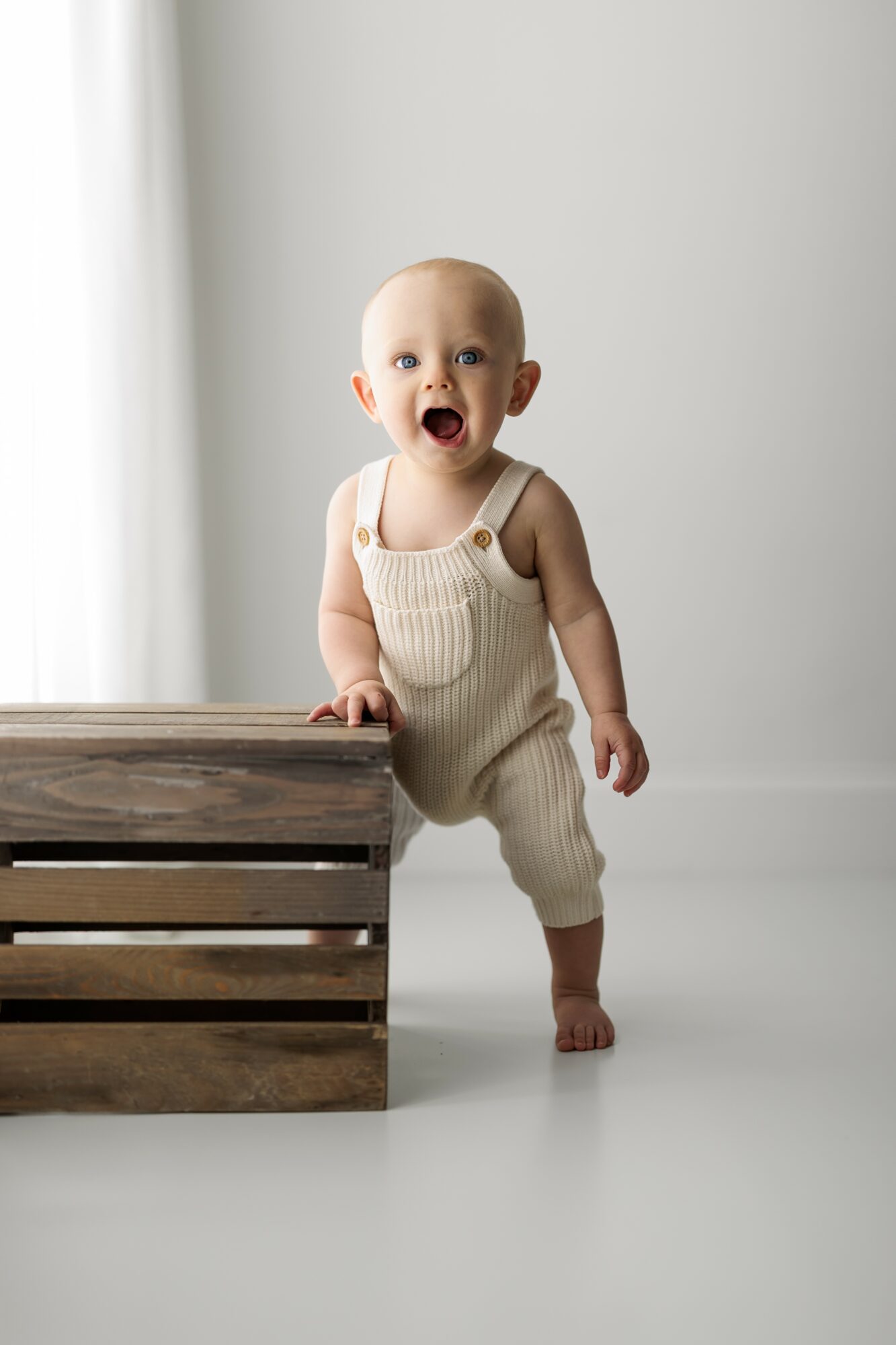 Baby standing next to a wooden crate, smiling with mouth open, in a bright room with white walls and curtains.