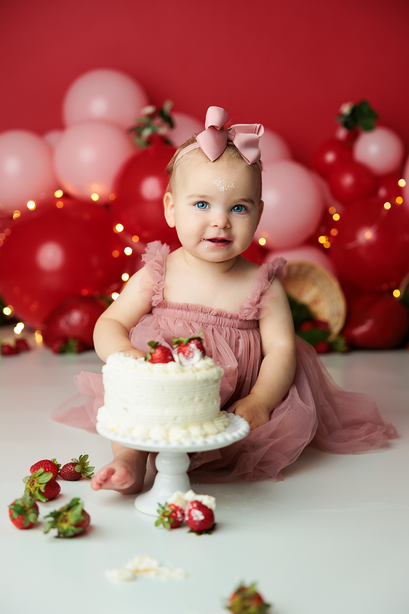 Baby girl in pink dress with a bow headband, sitting with a cake and strawberries, decorated with balloons and lights, smiling.