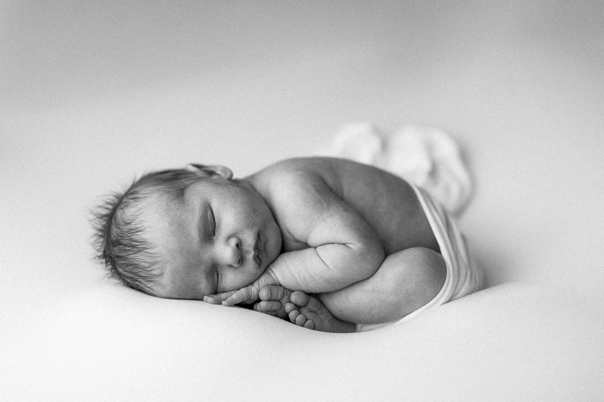 Sleeping newborn baby lying on side with hands near face, in black and white, soft background, peaceful scene.