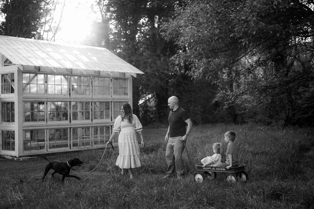 Two adults and two children outdoors near a greenhouse, with a dog on a leash and a wagon, trees in background.