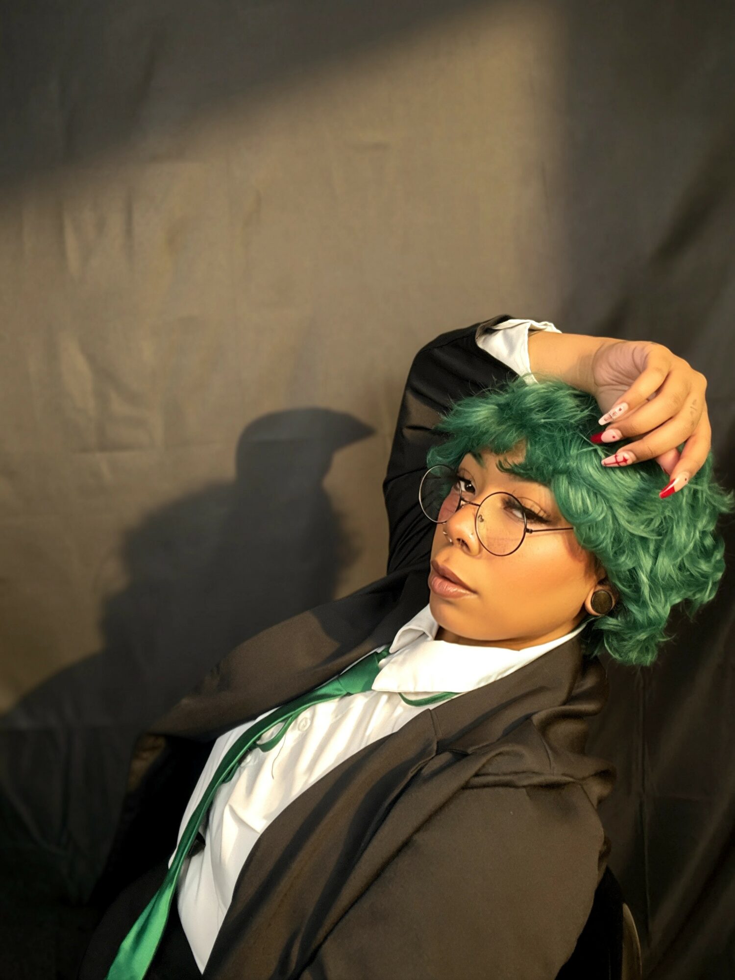 Person with green curly hair and glasses, wearing a black jacket and white shirt, resting head on hand against dark background.