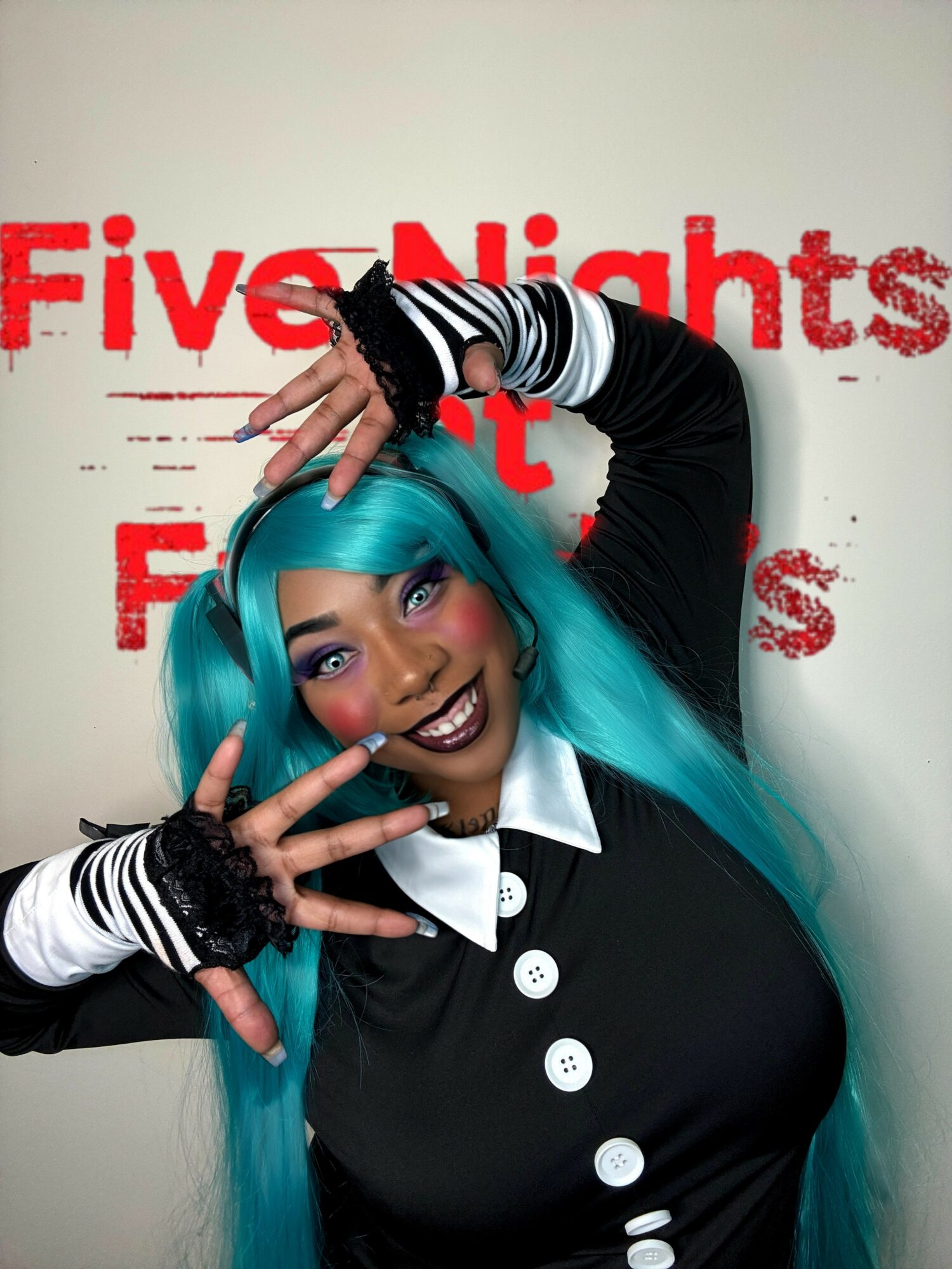 Person with long blue hair smiling, making a peace sign with both hands, standing in front of a wall with red text.