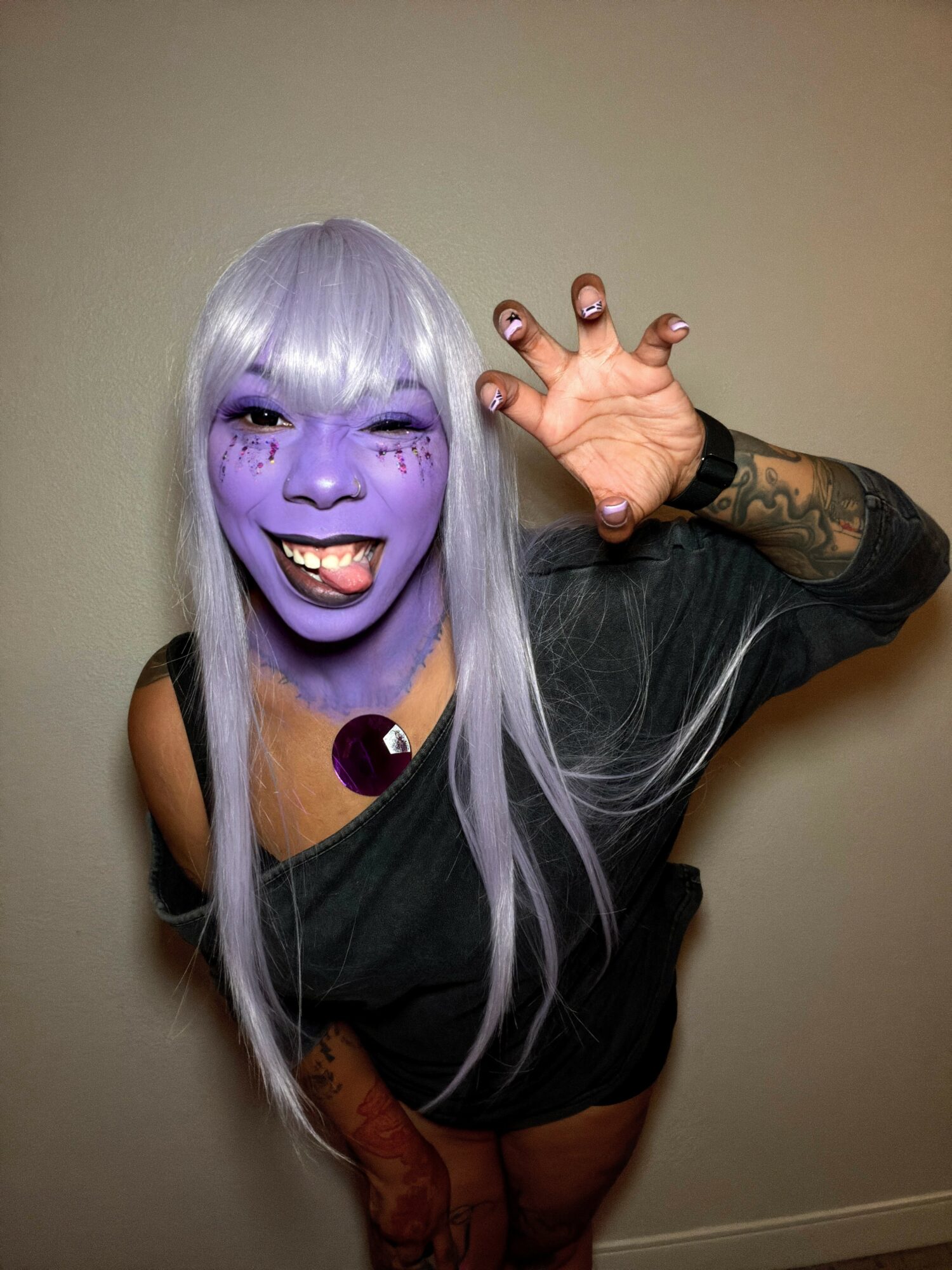 Person with purple face paint, silver hair, and a black top, reaching toward the camera with a playful expression.