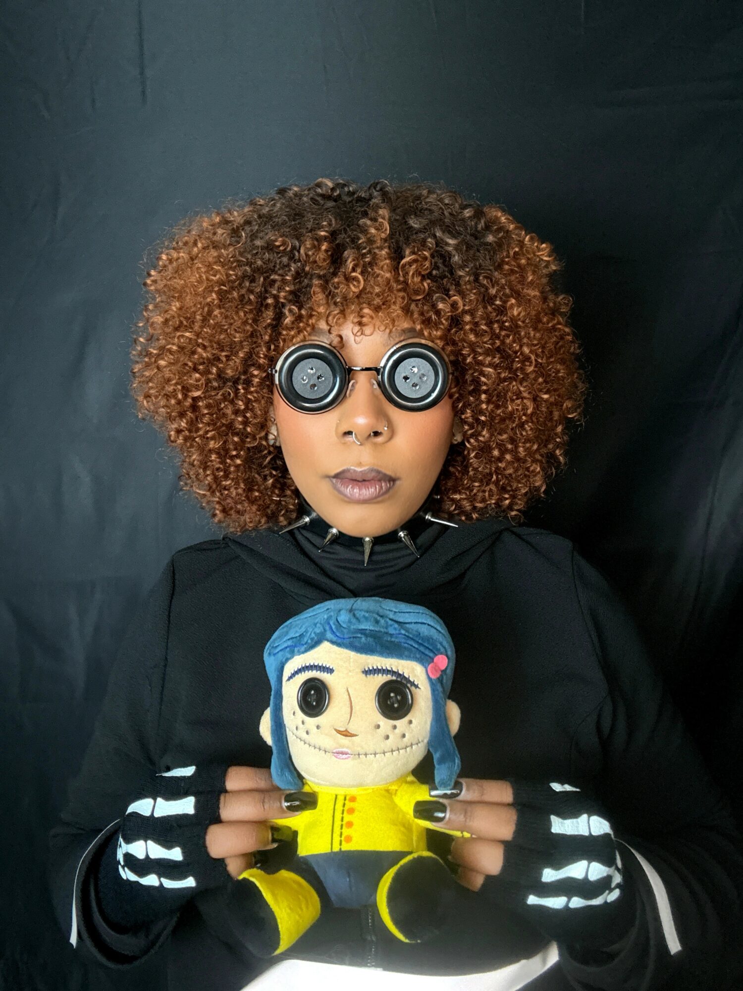 Person with curly hair wearing glasses and black clothing holding a plush doll with blue hair and yellow outfit, against a dark background.