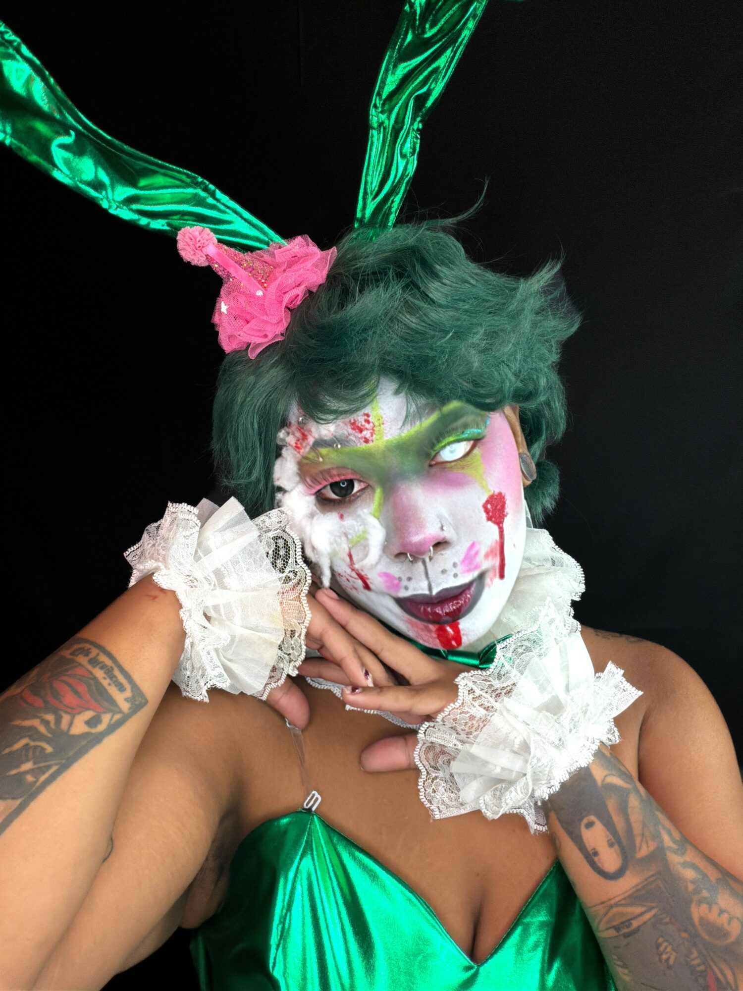 Person with green hair, colorful face makeup, and green bunny ears, wearing white lace cuffs, posing with hands near face.