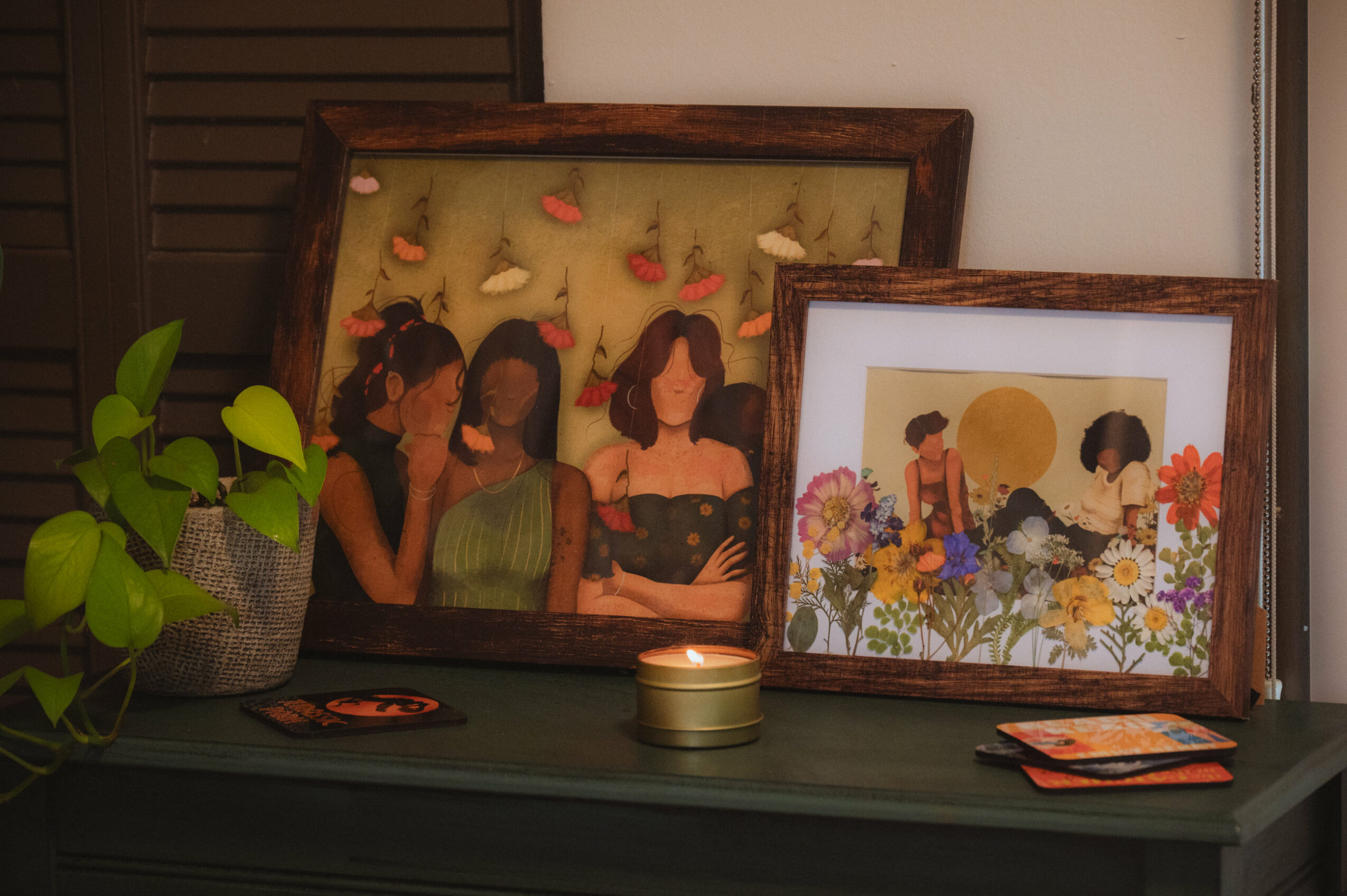 Two framed paintings on a dark surface, a potted plant, a small gold container, and some cards or photographs, indoors.