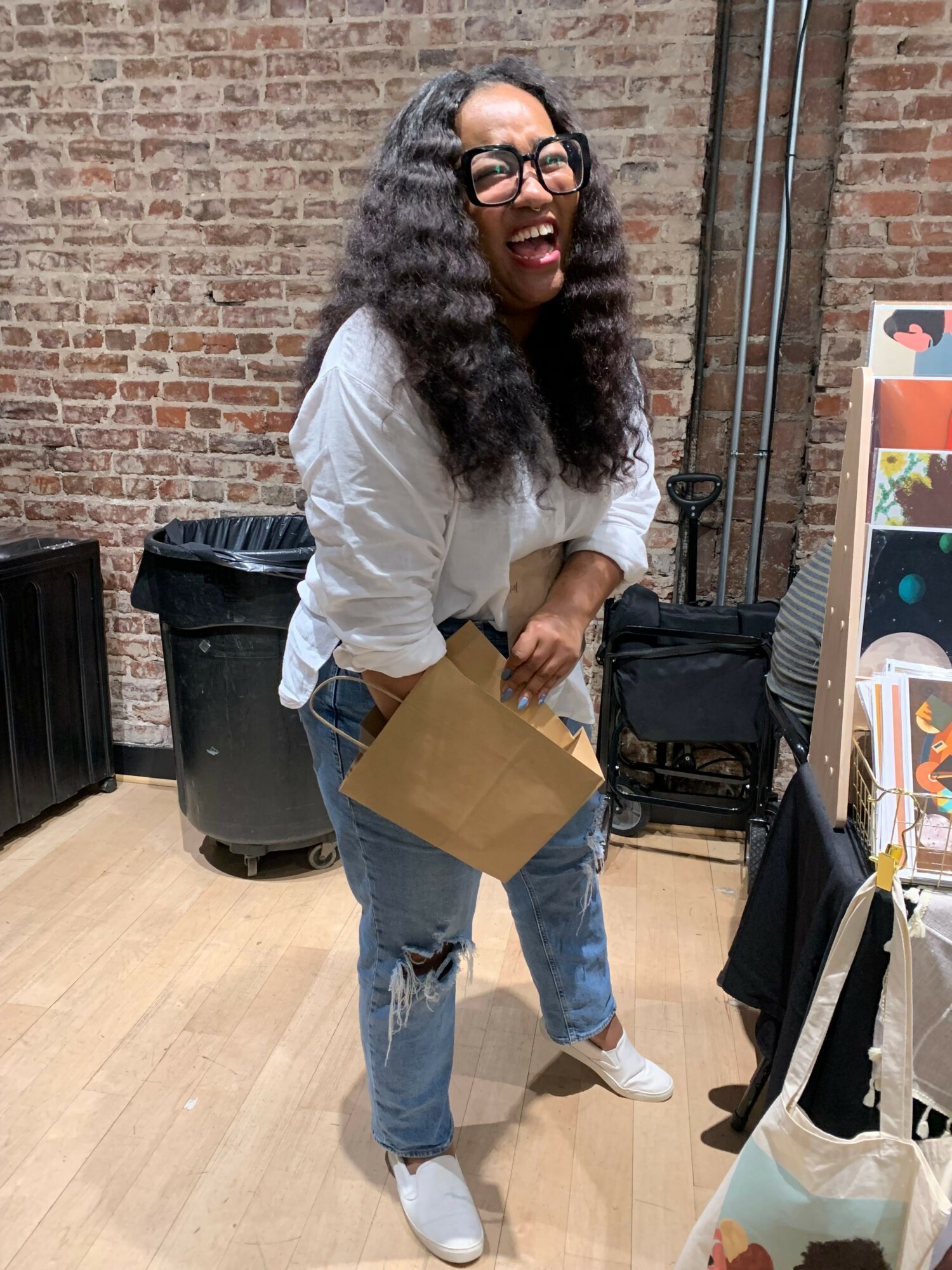 Woman with curly hair, glasses, white shirt, and jeans holding a cardboard box, smiling, standing indoors near a brick wall.