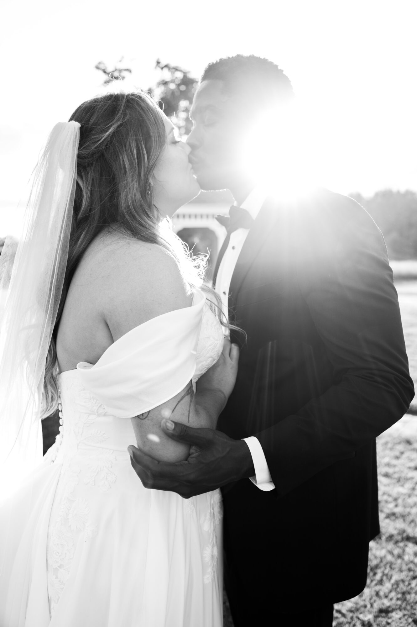 A bride and groom face each other outdoors, with sunlight behind them, holding hands and about to kiss, in black and white.