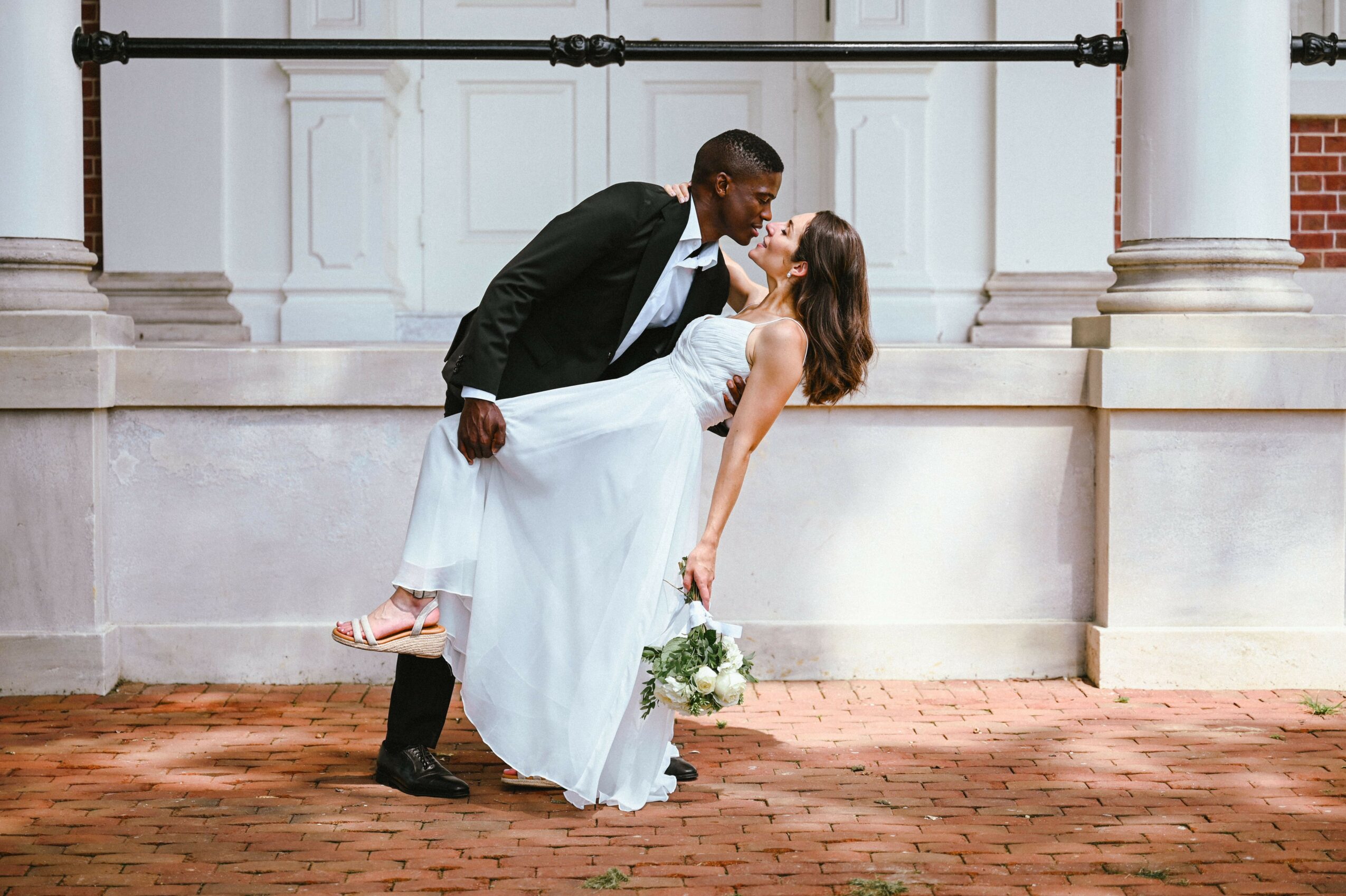 A couple in wedding attire sharing a kiss outdoors, with the groom dipping the bride, holding a bouquet, in front of a building with columns.