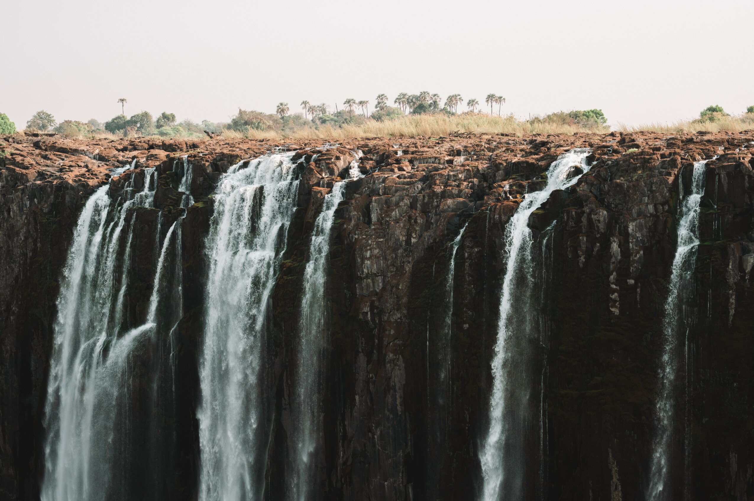 Waterfalls cascading over a rocky cliff with a distant tree line and cloudy sky in the background.