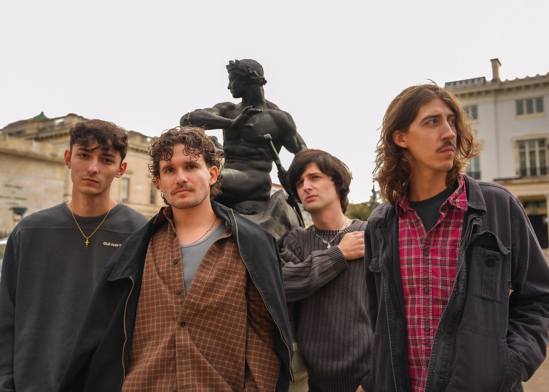 Four young men stand in front of a statue in an urban setting, with buildings in the background.