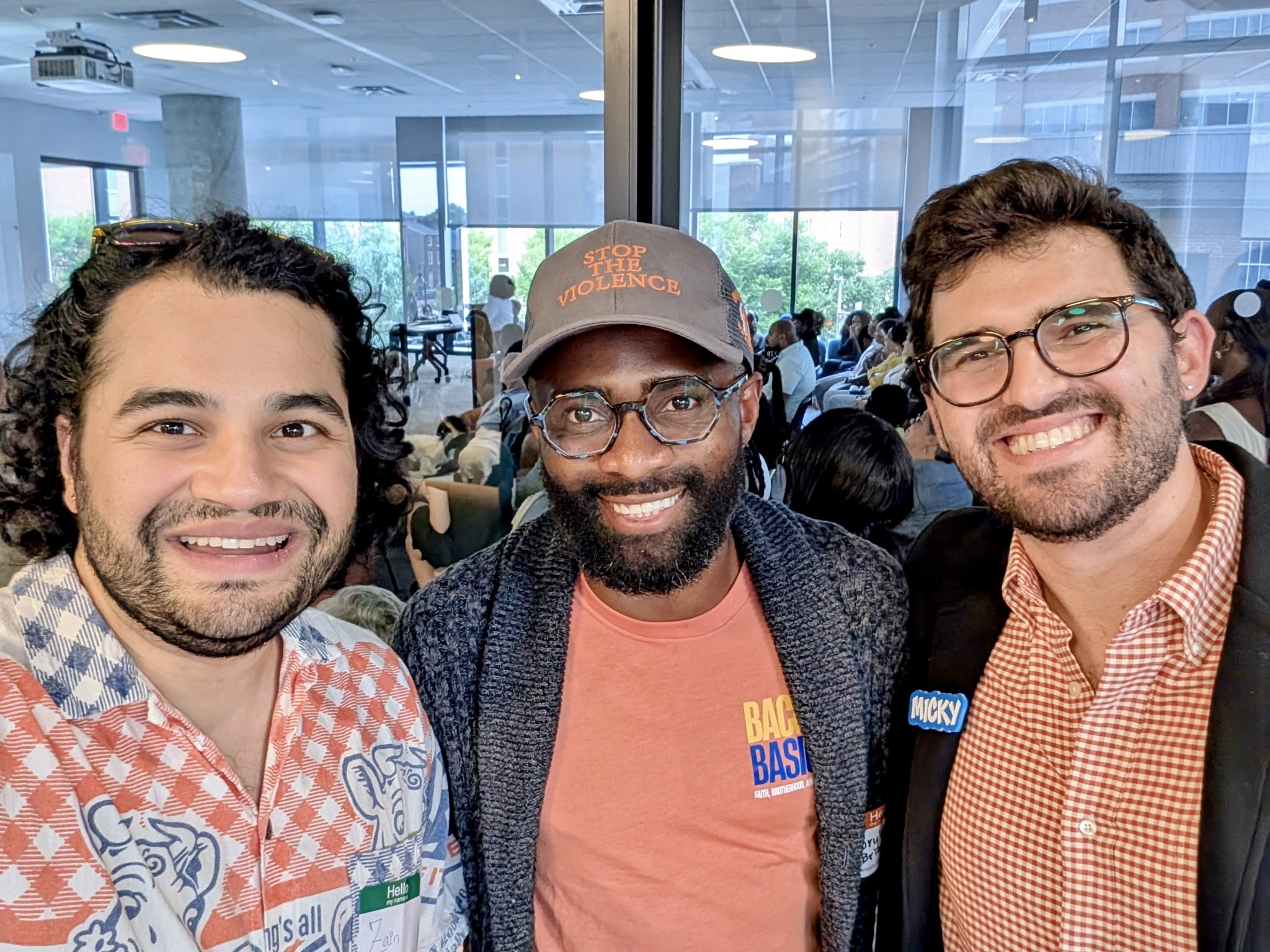 Three smiling men standing together indoors, with a crowd and large windows in the background.