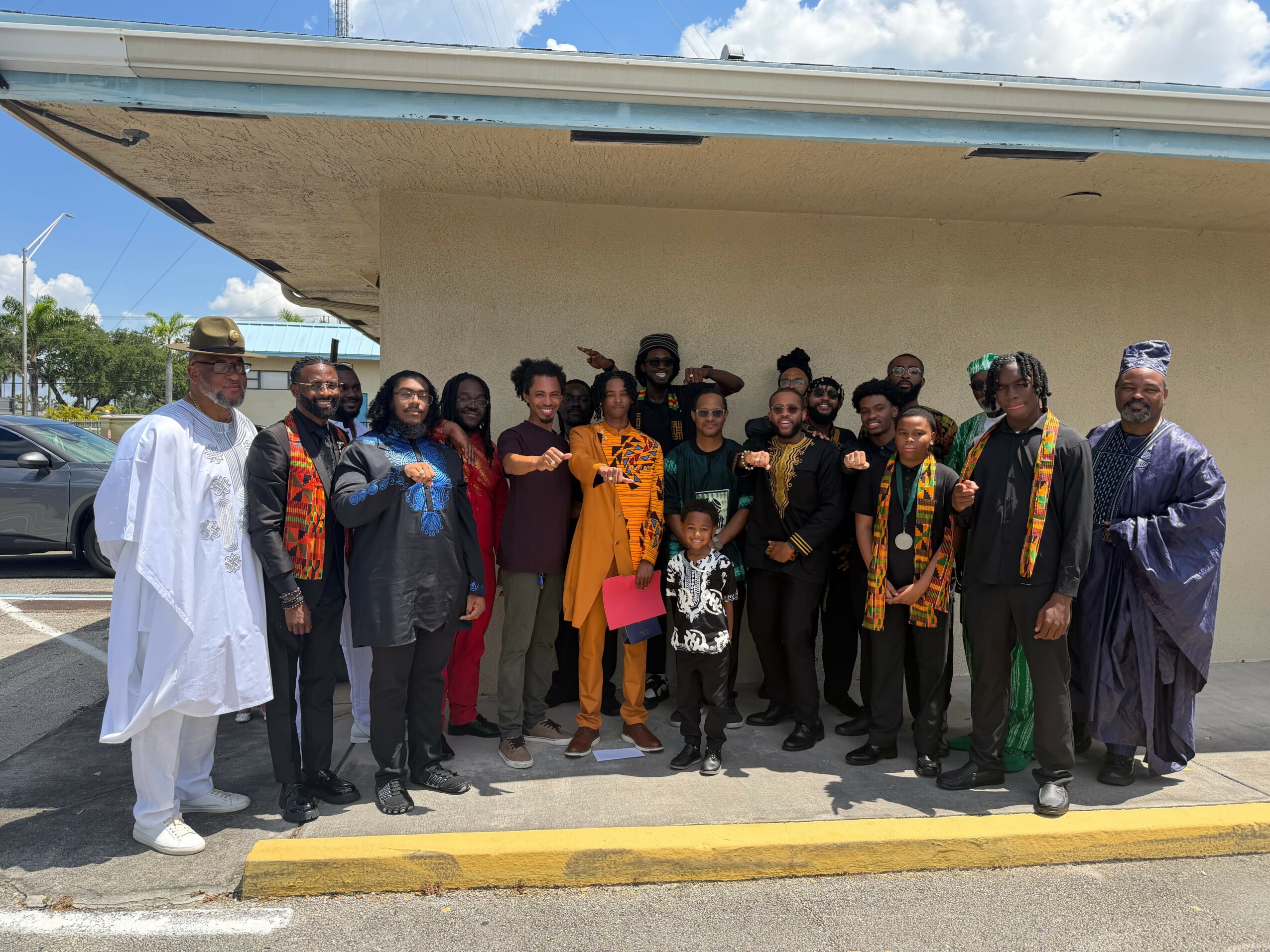 Group of people standing outside a building, some wearing traditional clothing, under a blue sky with clouds.