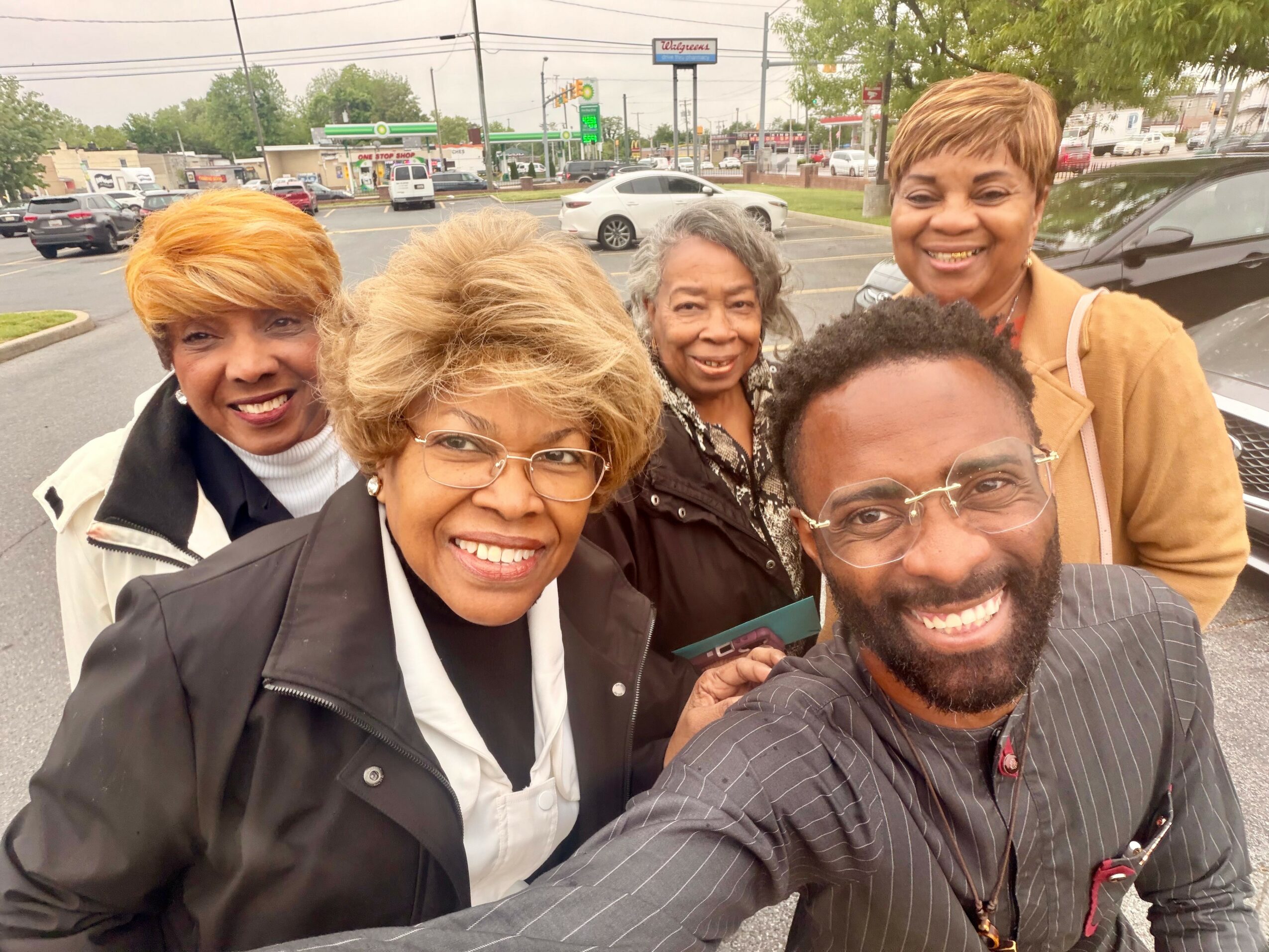 Group of five smiling people standing outdoors in a parking lot, with cars and trees in the background.