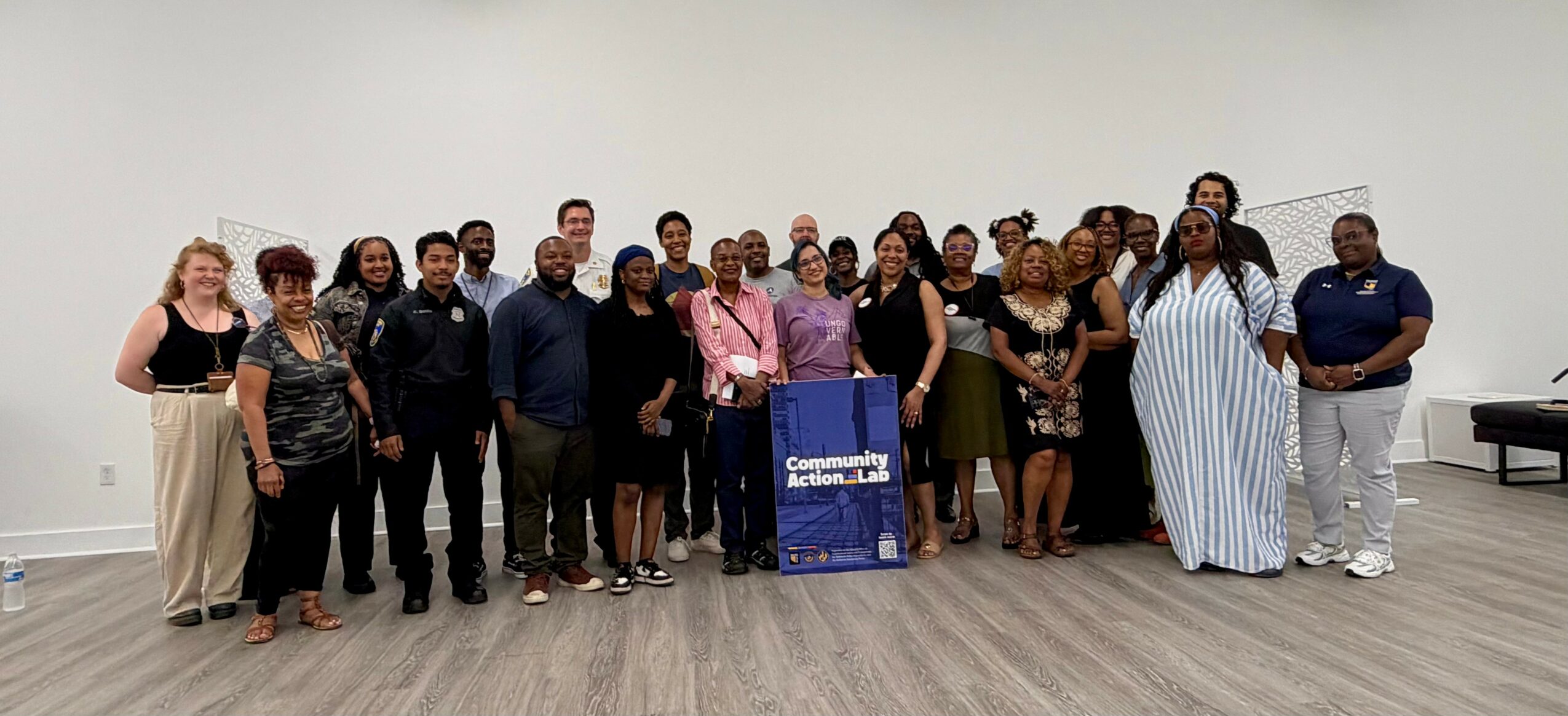 Group of people standing in a room with a sign, some smiling, some with neutral expressions, diverse clothing styles, plain white wall background.