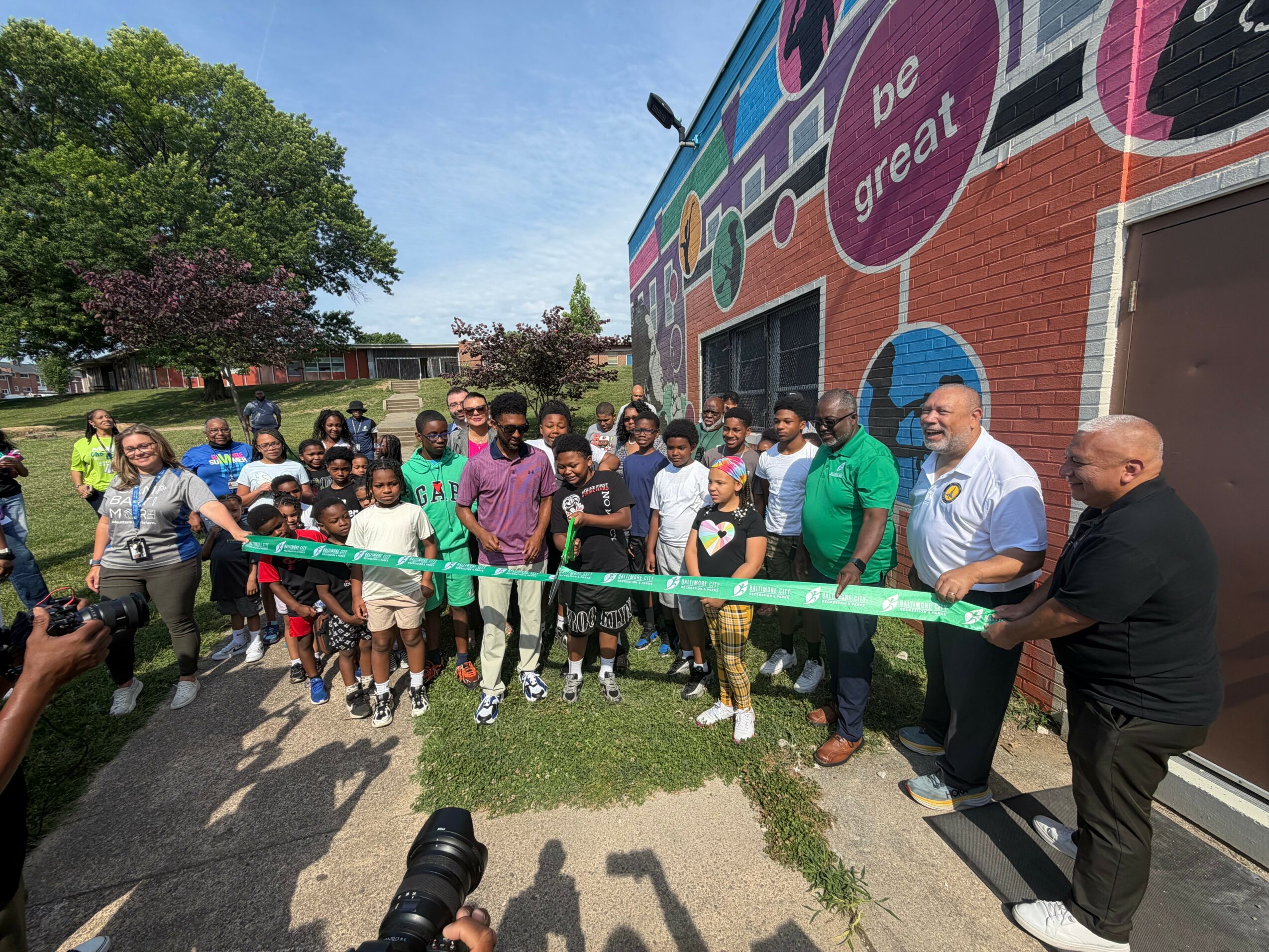 Group of people gathered outside a building with a colorful mural, some holding a green ribbon for a ribbon-cutting event.