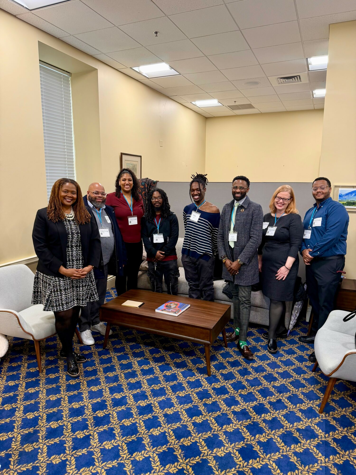 Group of eight diverse people standing in a room with beige walls and blue patterned carpet, smiling for a photo.