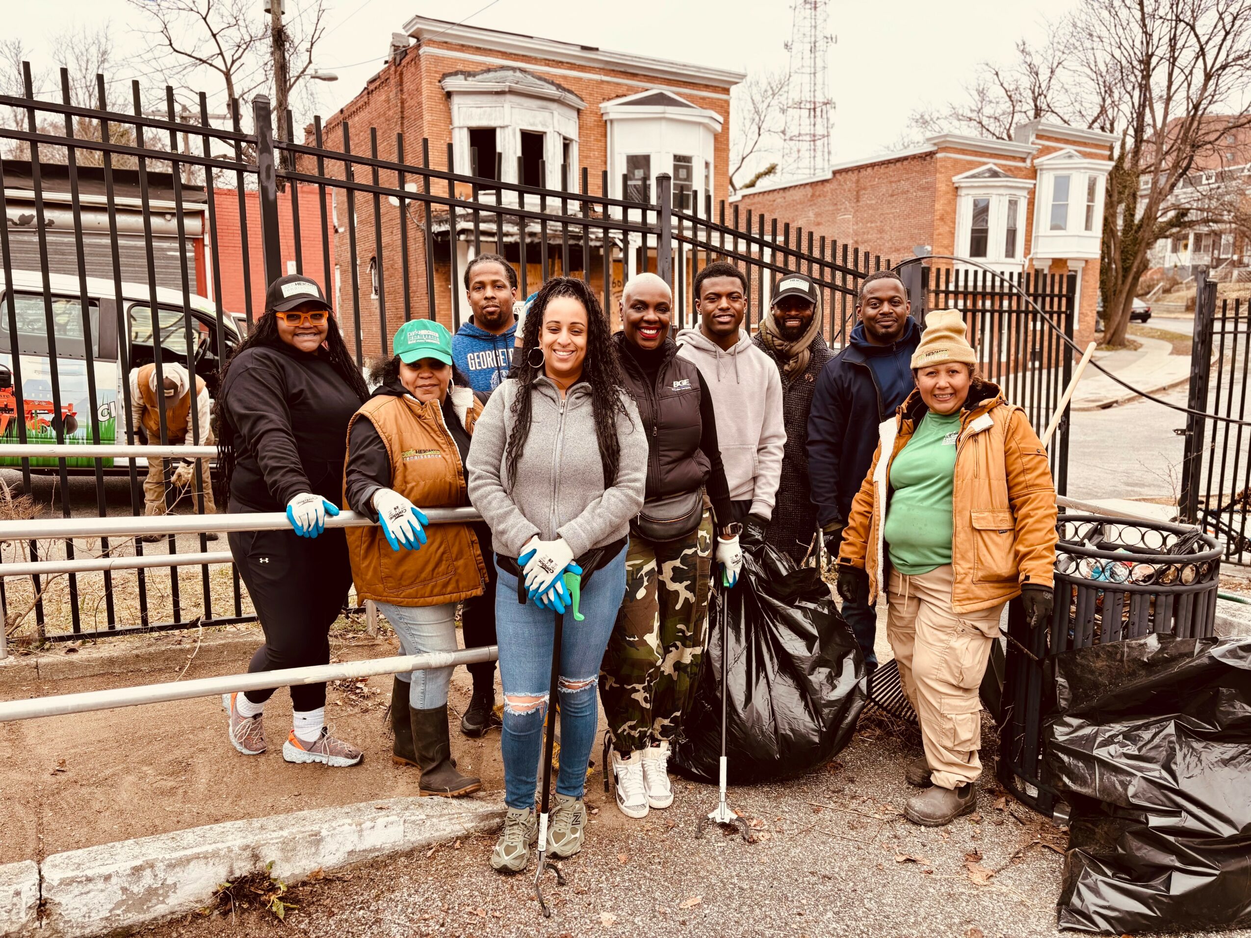 Group of ten diverse people standing outdoors behind a black fence, some holding trash bags, participating in a cleanup event.