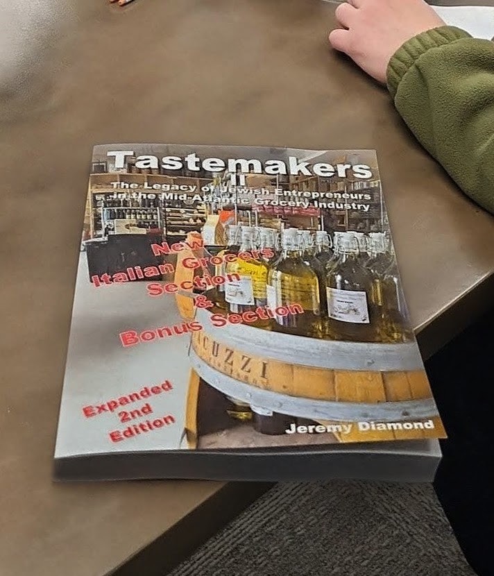 Book titled 'Tastemakers' with bottles of alcohol on a bar counter, partially visible person's arm in green sleeve, on a brown table.